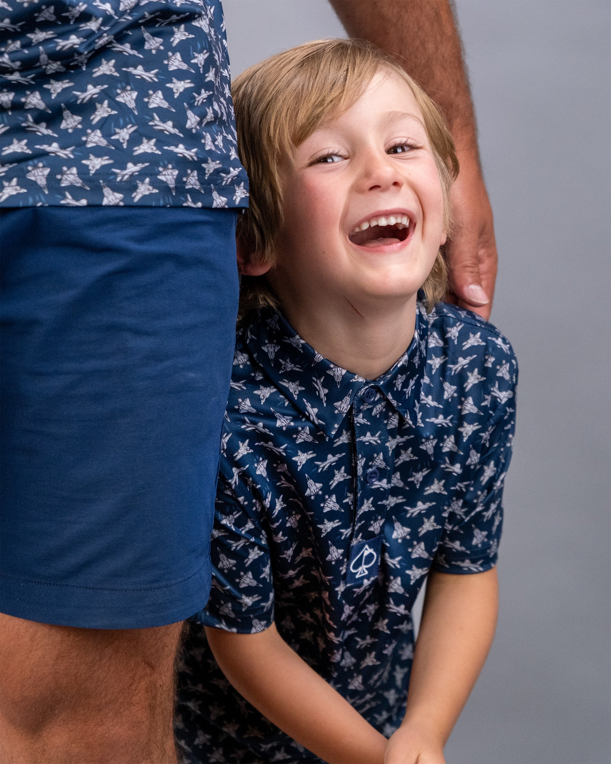 A young boy with blonde hair smiling and laughing, wearing a navy shirt with a bird pattern, standing next to an adult.