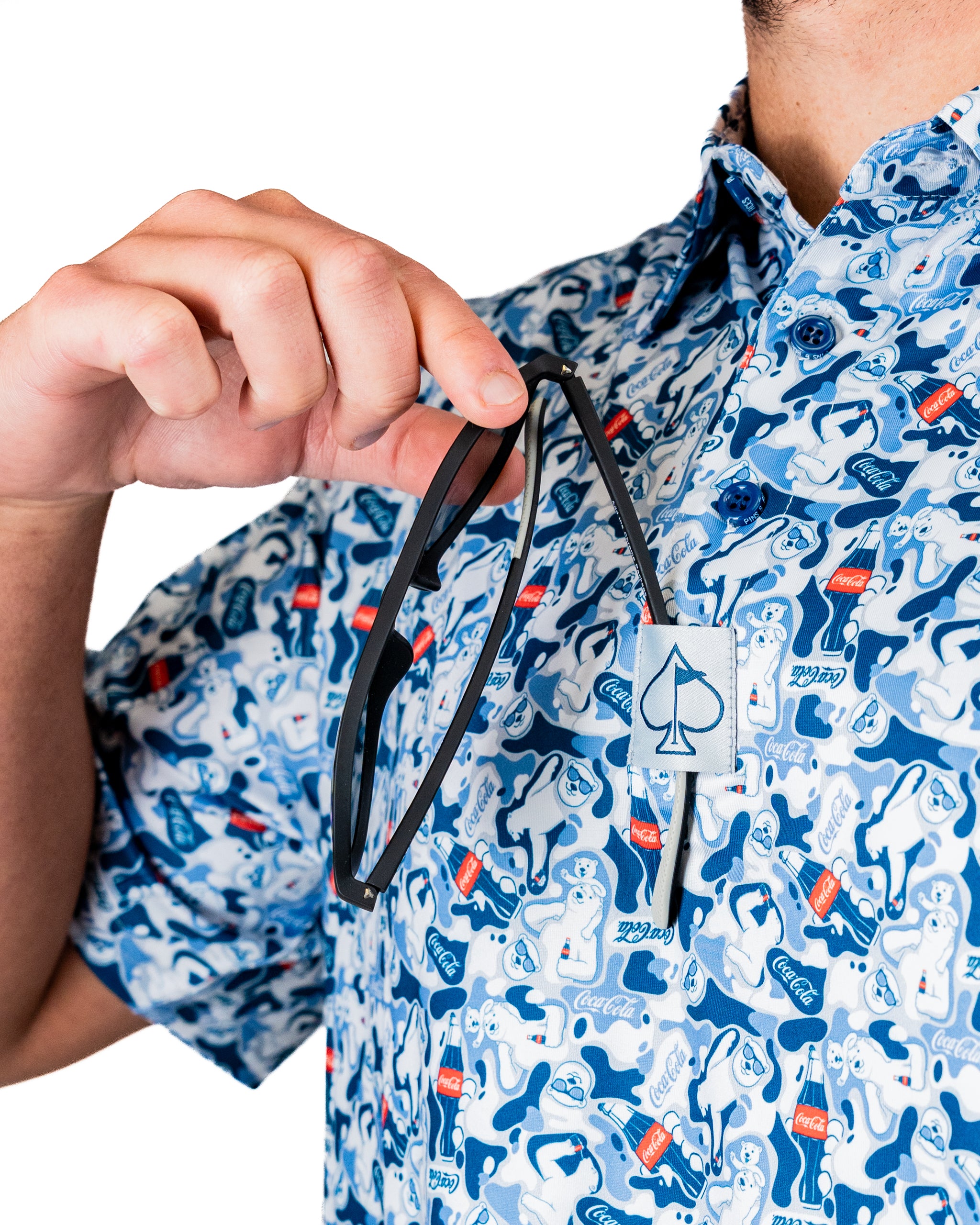 A person holds a black golf headcover while wearing a blue patterned shirt featuring Coca-Cola designs.