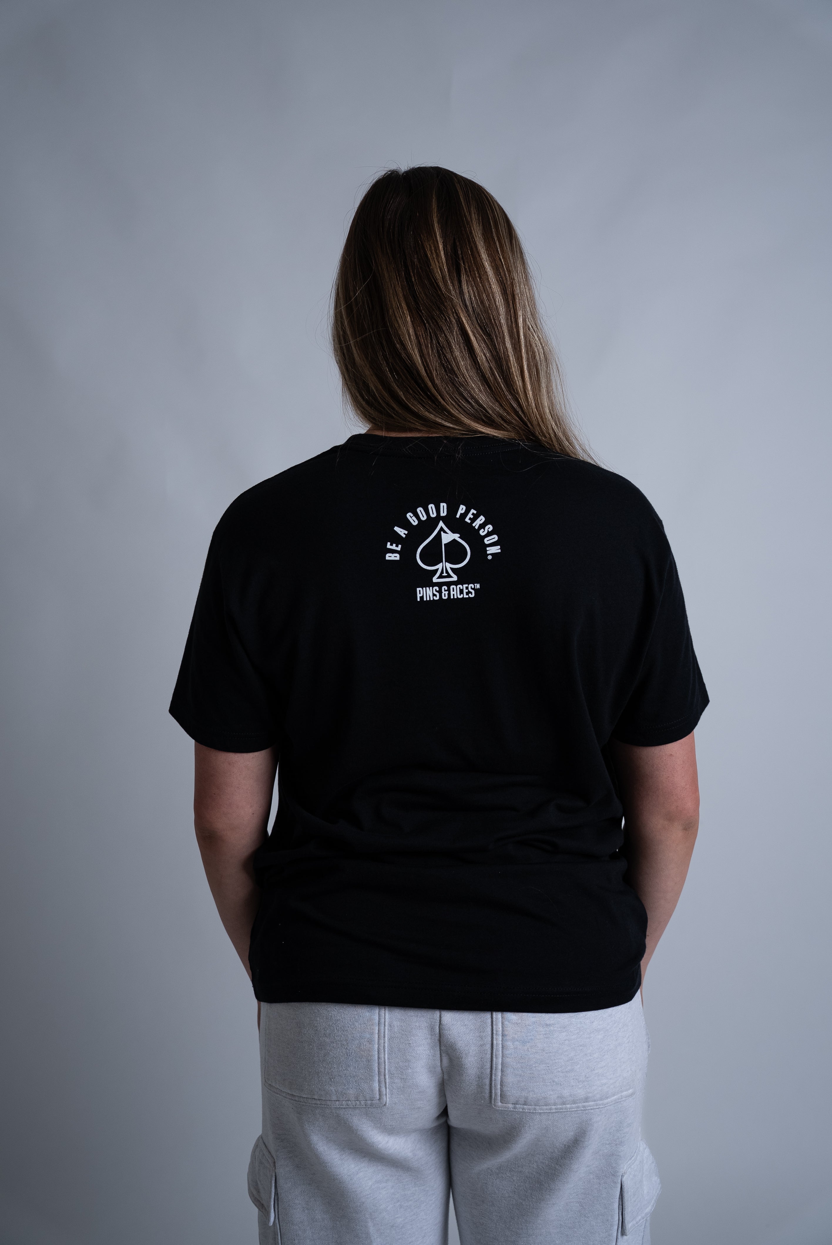 Black Be A Good Person X Pins and Aces signature t-shirt featuring a logo on the back, modeled by a person from behind.