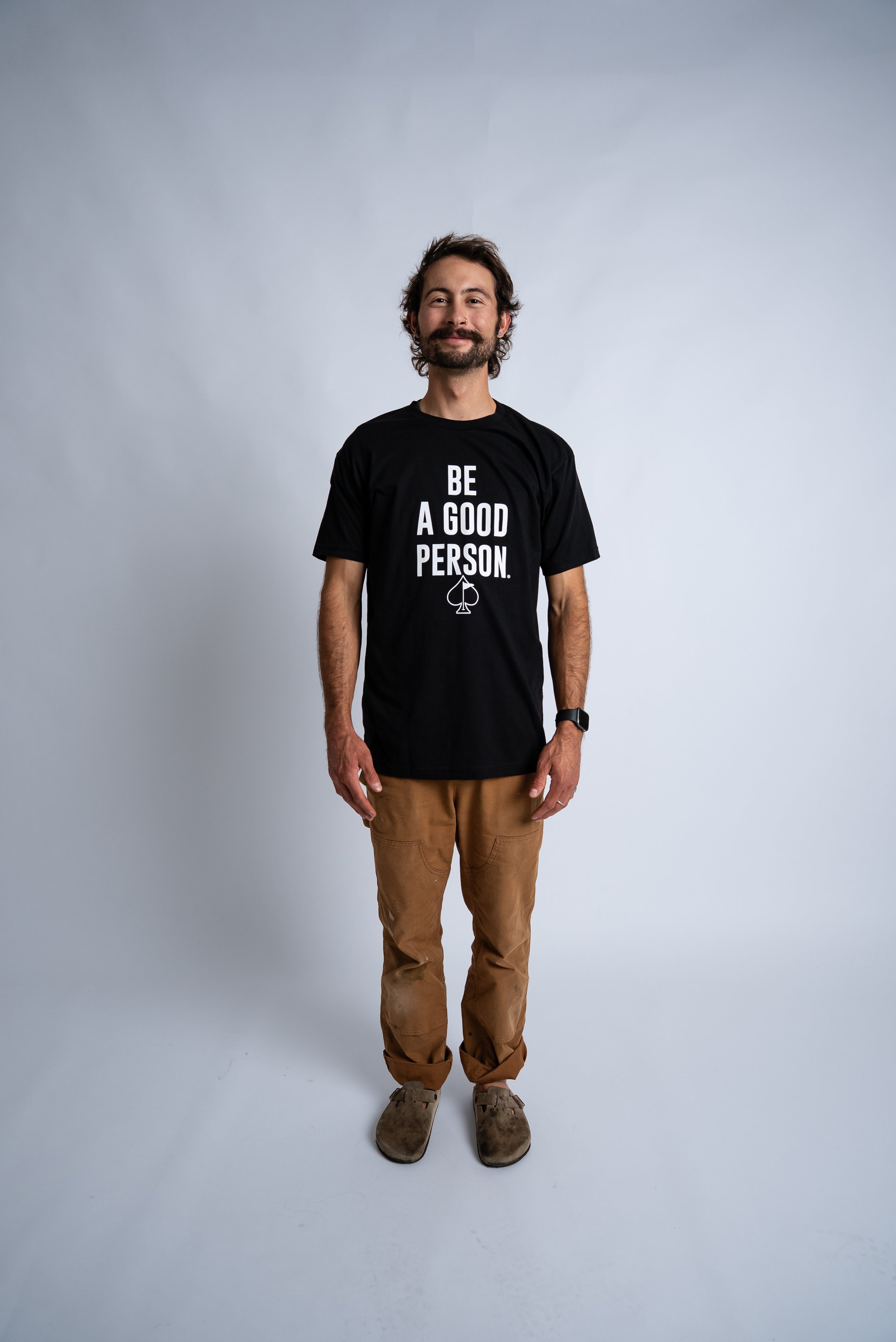 Man standing in a black "Be A Good Person" t-shirt, smiling, wearing khaki pants and casual shoes.