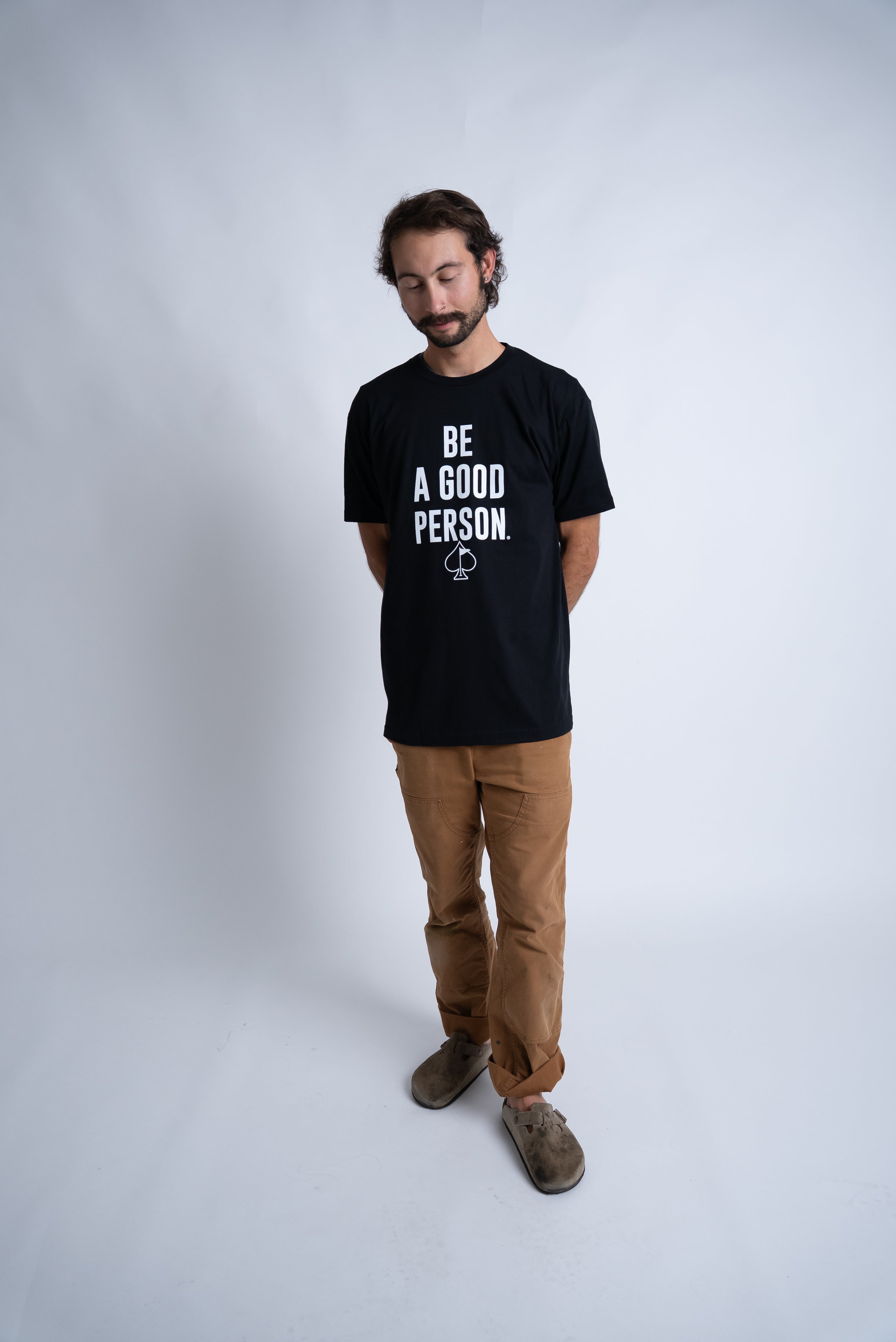 Model wearing a black "Be A Good Person" t-shirt from Pins and Aces, paired with brown pants and standing against a plain background.