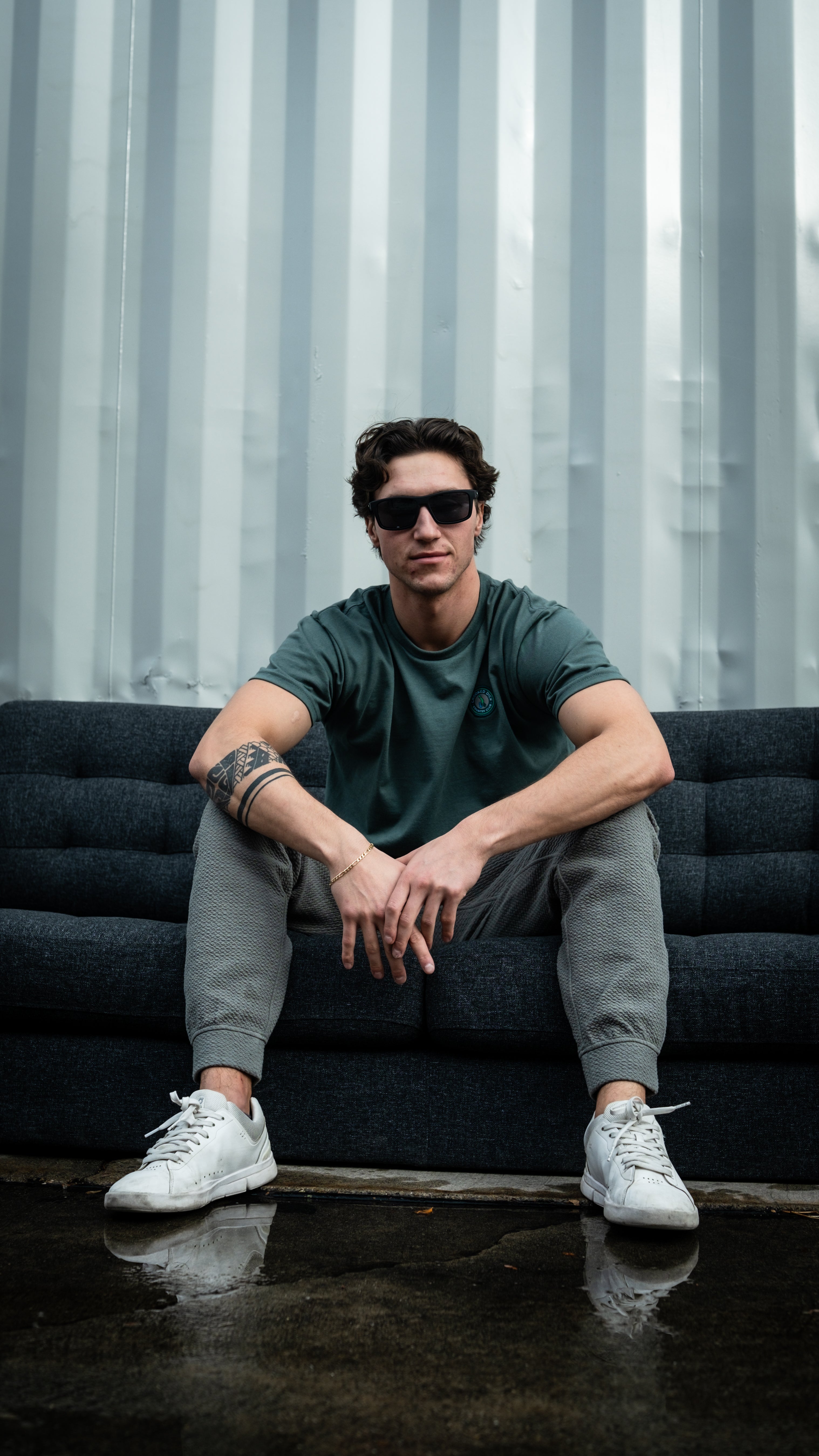 Stylish man in black sunglasses, green t-shirt, grey joggers, and white sneakers sitting on a dark sofa against a metallic...