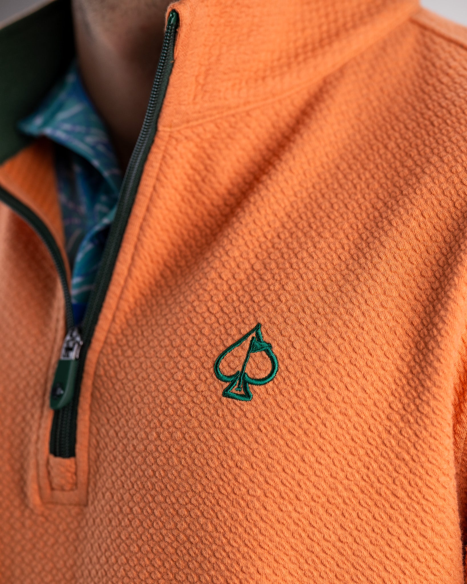Close-up of an orange golf jacket with a green embroidered spade logo, black zipper, and layered patterned shirt underneath.