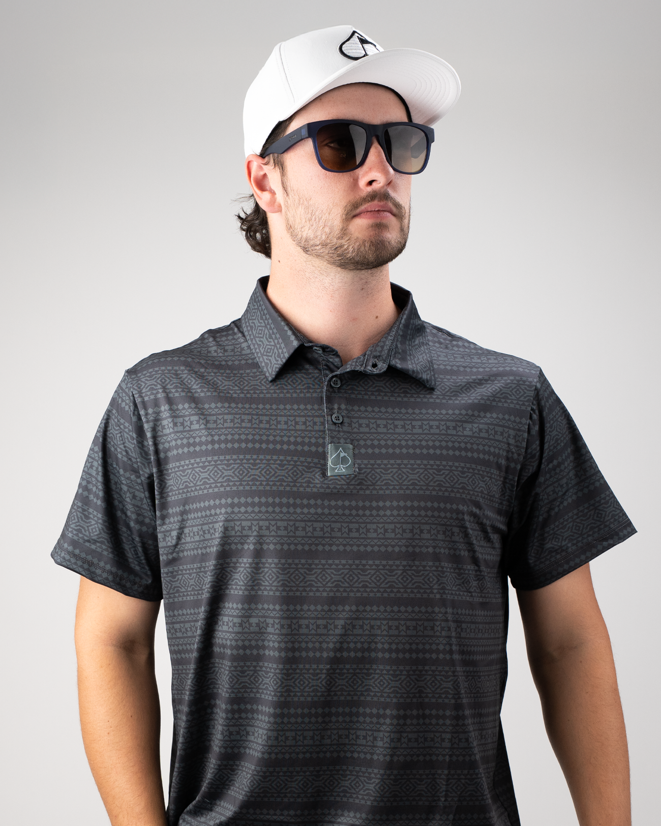 Man wearing a Desert Geo golf polo shirt with a geometric pattern, sunglasses, and a white cap, posing confidently.