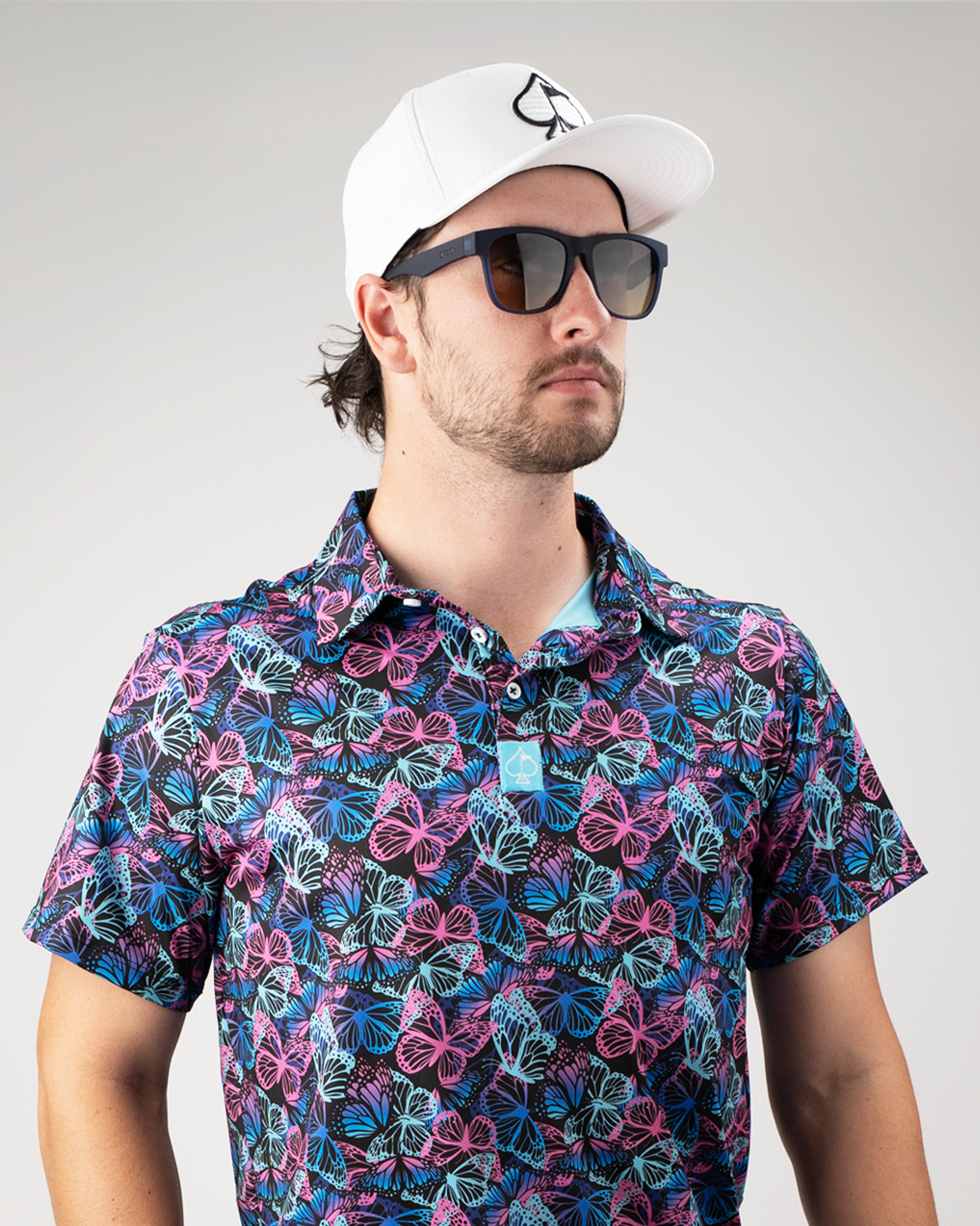 Man wearing a vibrant butterfly-patterned polo shirt and sunglasses, paired with a white cap, showcasing bold golf style.
