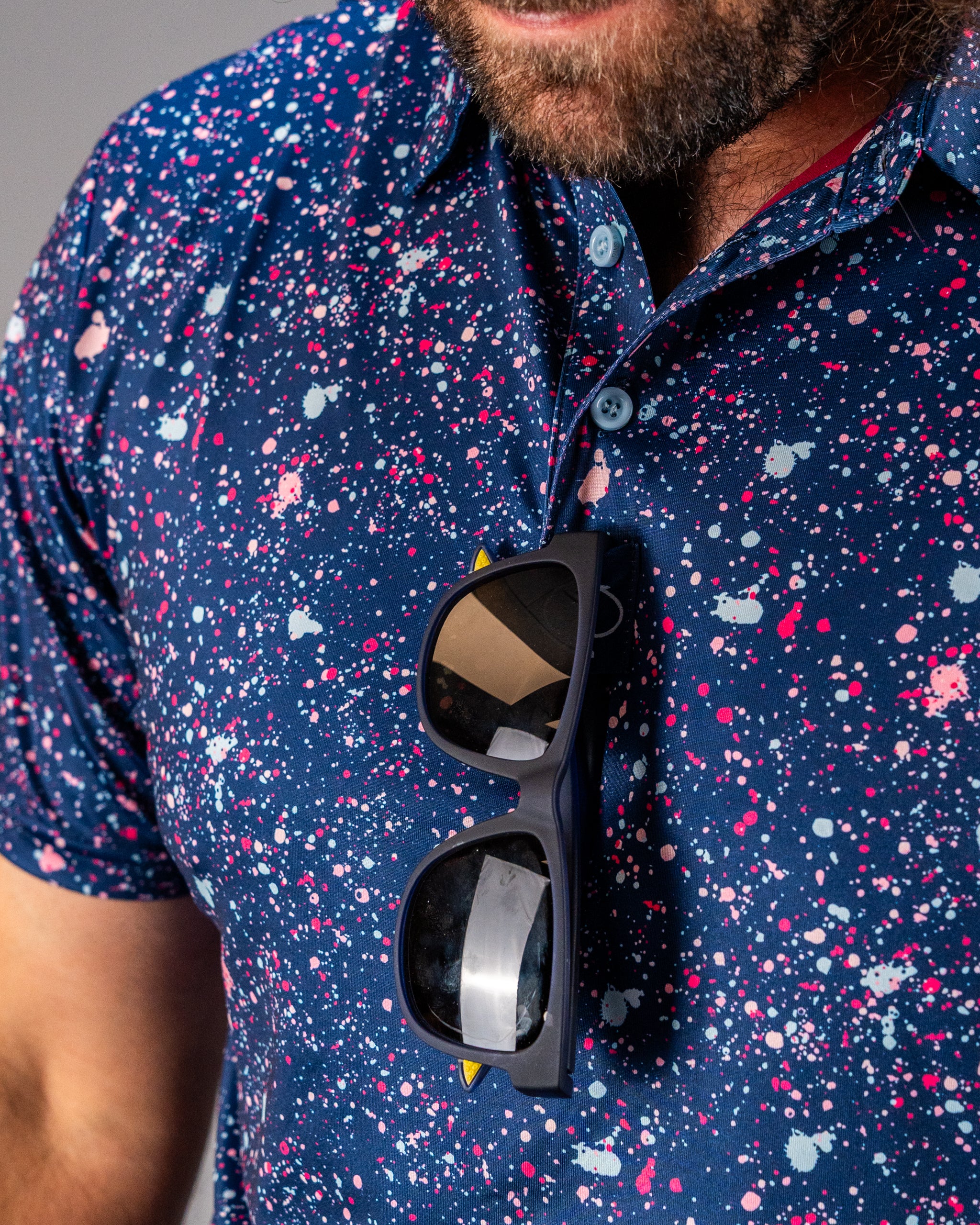 Men's blue splatter golf shirt featuring vibrant paint splashes, paired with stylish sunglasses hanging from the collar.