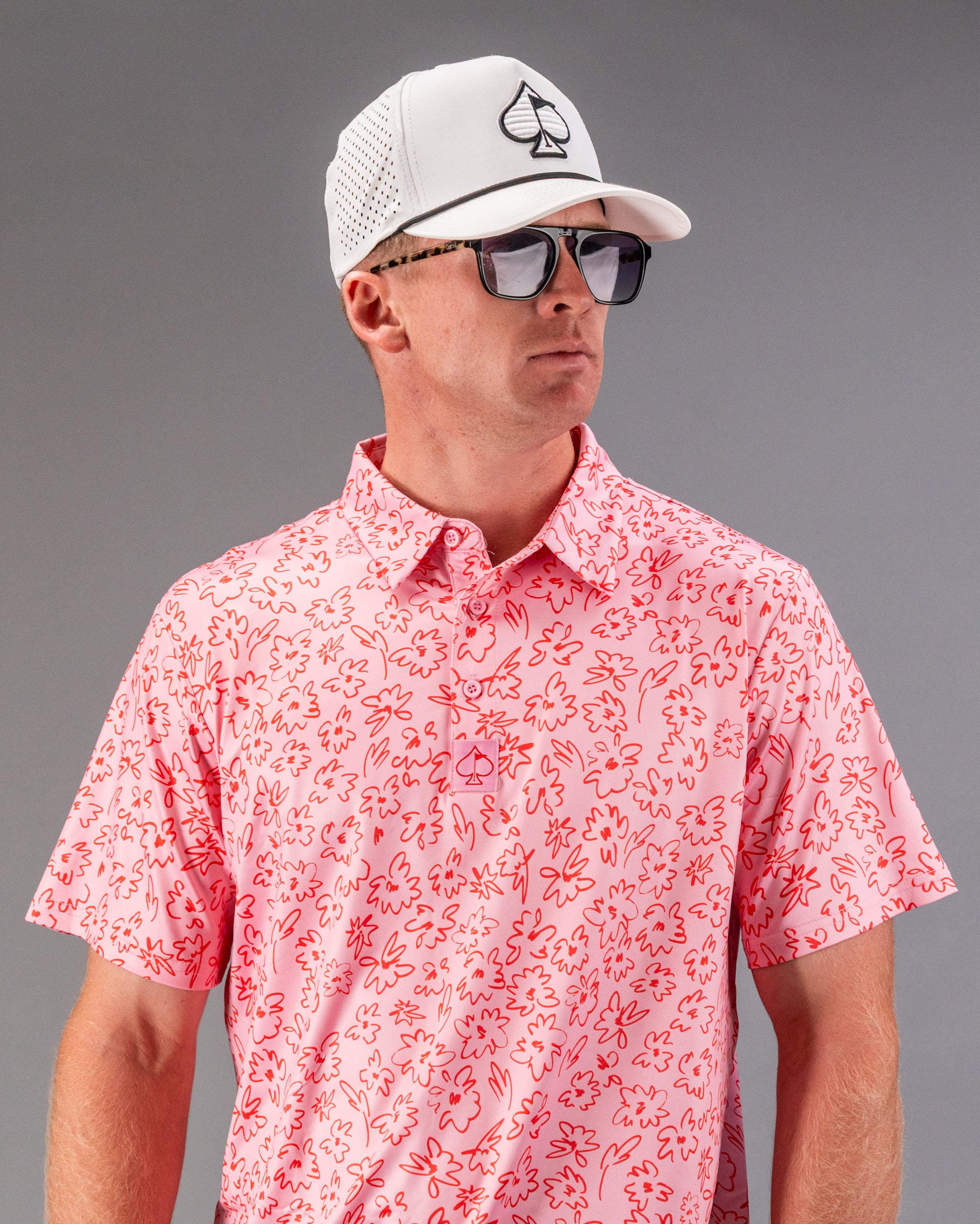 Men's pink golf shirt featuring a playful flower doodle pattern, paired with a white cap and sunglasses for a stylish look.
