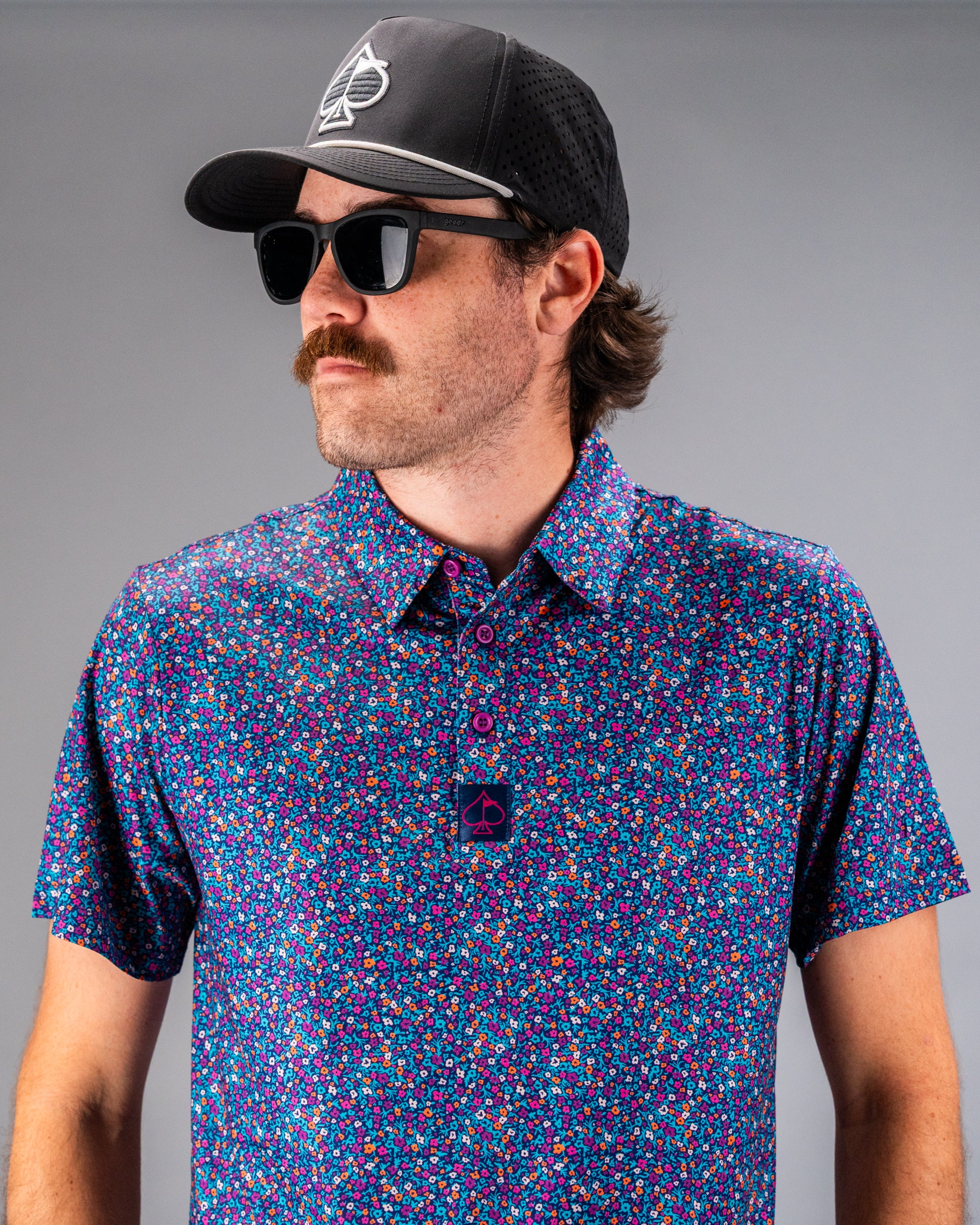 Man wearing a vibrant lavender floral golf shirt with a black cap and sunglasses, showcasing bold style on the course.