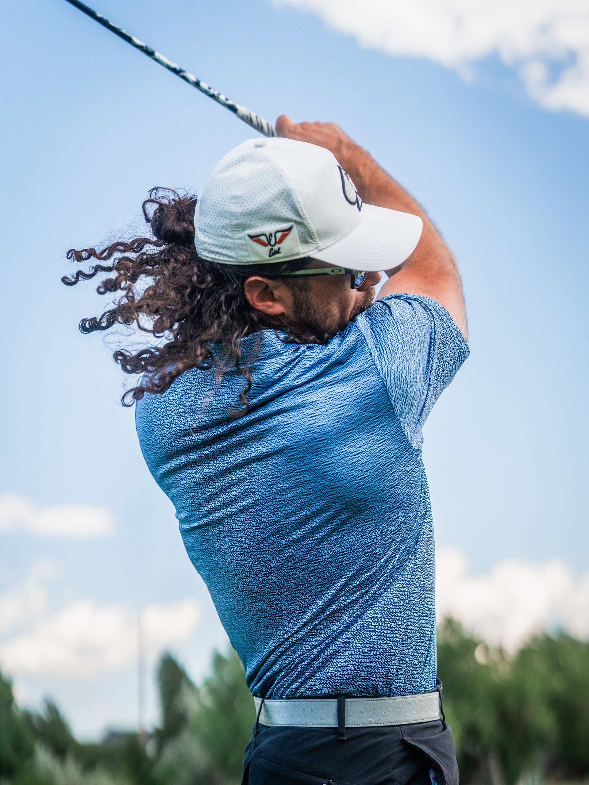A golfer swings a club, showcasing the Squiggle Blue golf shirt with a textured pattern against a sunny sky.
