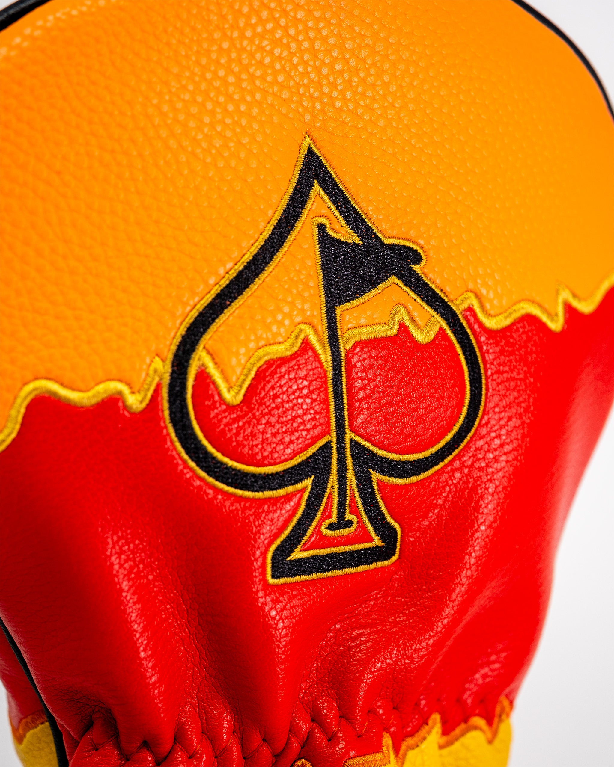 Close-up of a golf driver cover featuring a fiery red and yellow design with a black spade logo embroidered in the center.