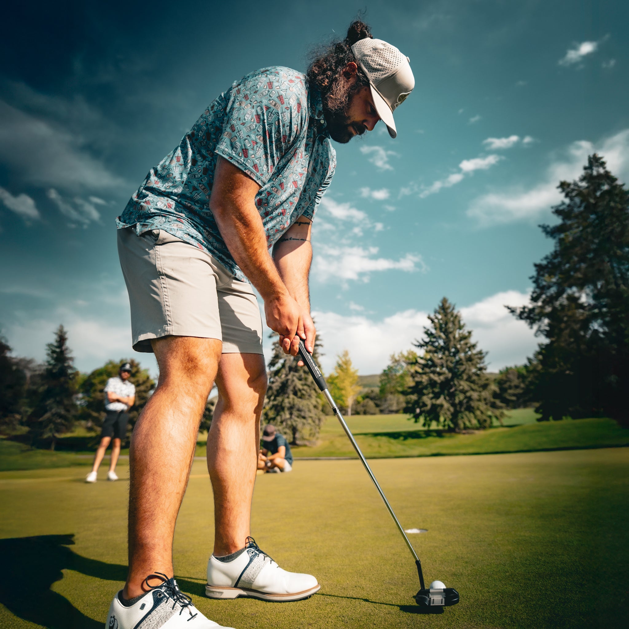 Golf player in colorful shirt and white shoes preparing to putt on the green, with trees and cloudy sky in the background.