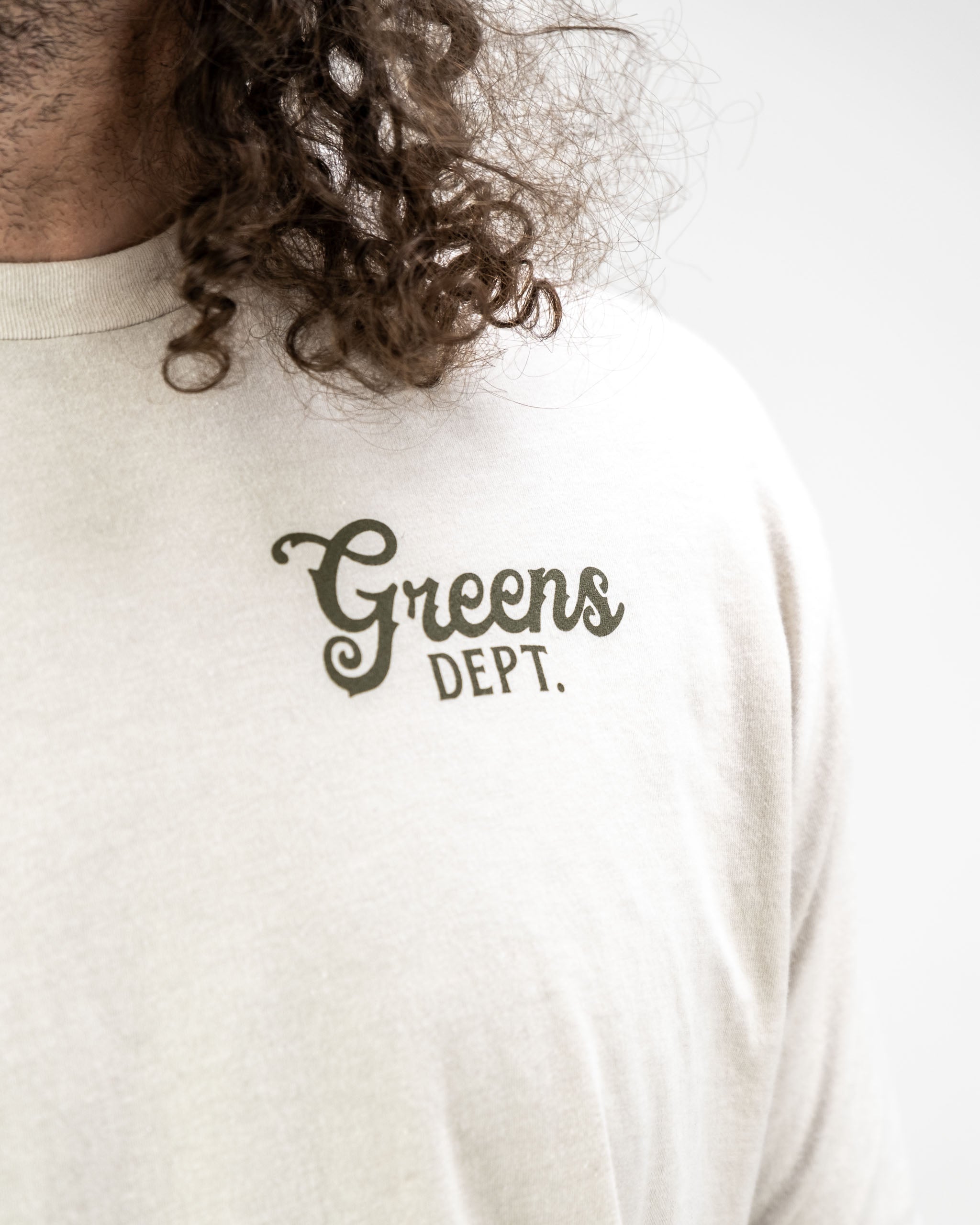 Light-colored tee shirt featuring "Greens Dept." text in a stylish font, designed for golf enthusiasts.