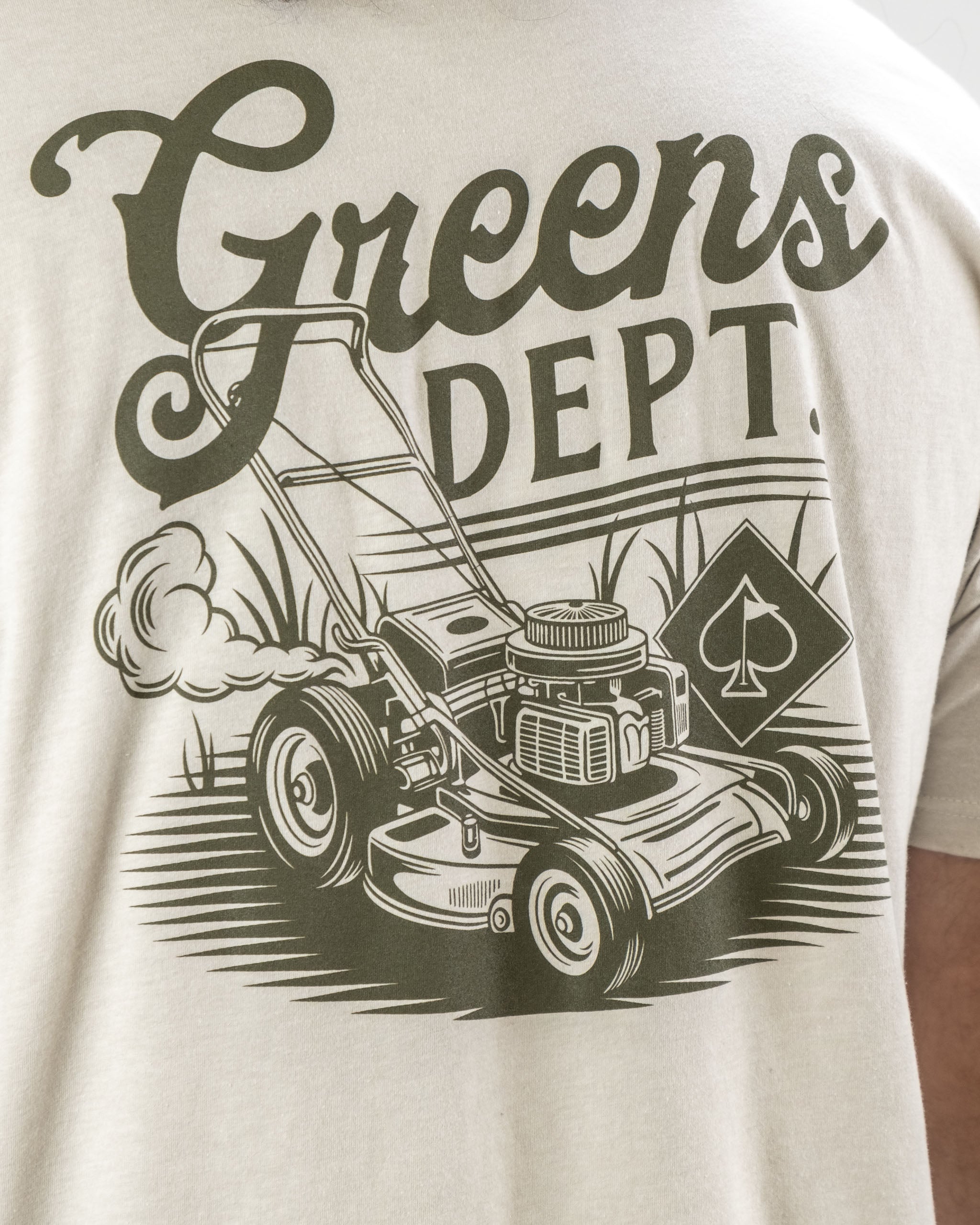 Beige tee shirt featuring a vintage lawnmower graphic with "Greens Dept" text, perfect for golf enthusiasts.