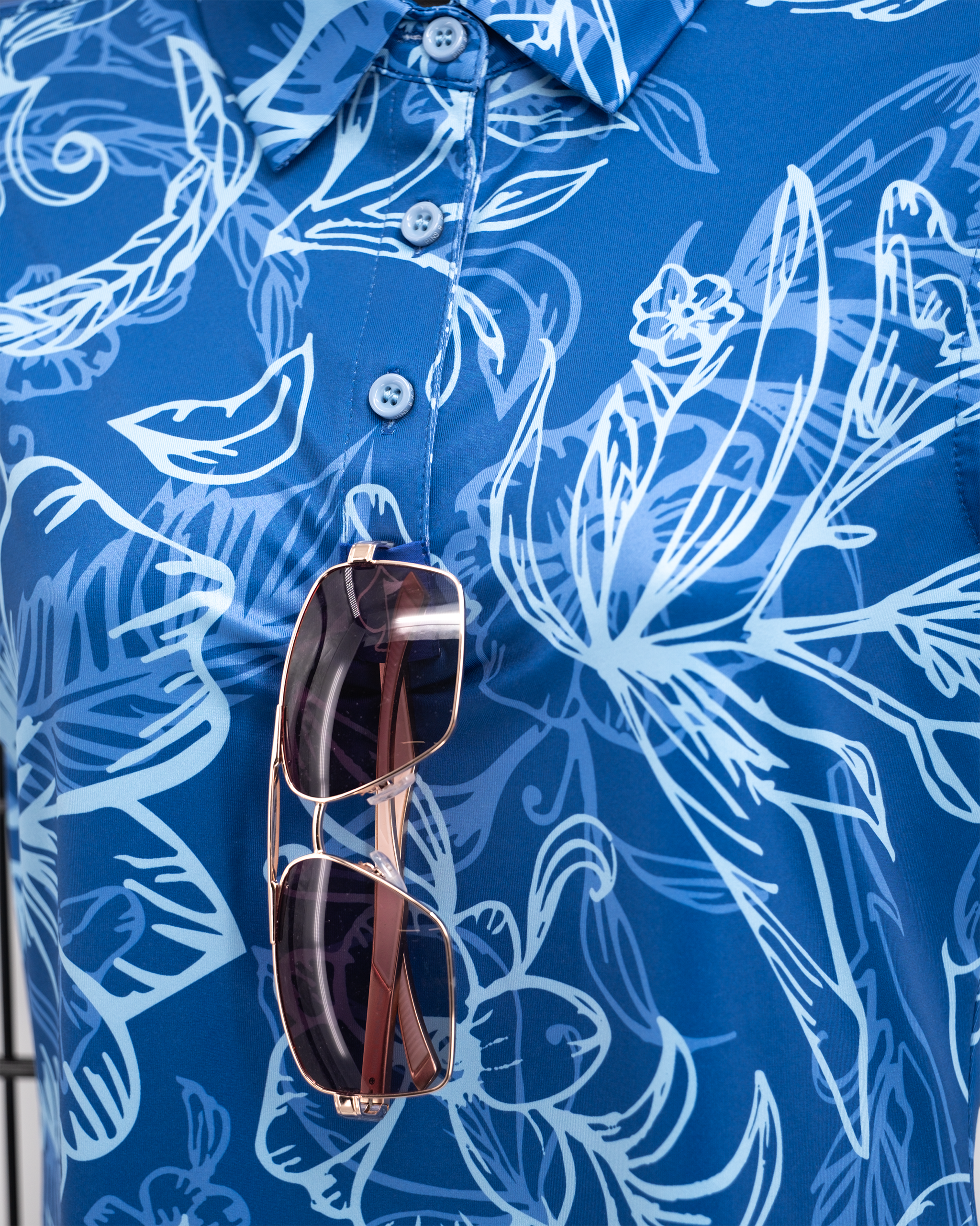 Blue floral patterned golf shirt with white outlines, featuring a collar, buttons, and sunglasses hanging from the placket.