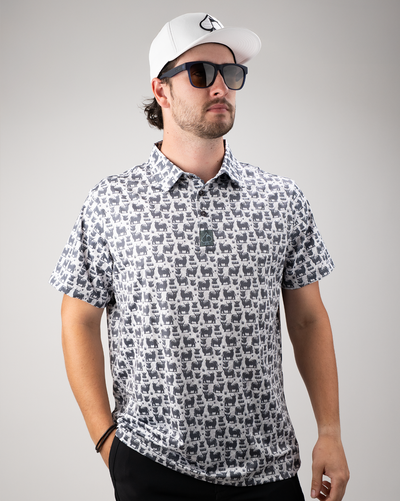 Man wearing a short-sleeve golf shirt featuring a Highland cow pattern, paired with sunglasses and a white cap.