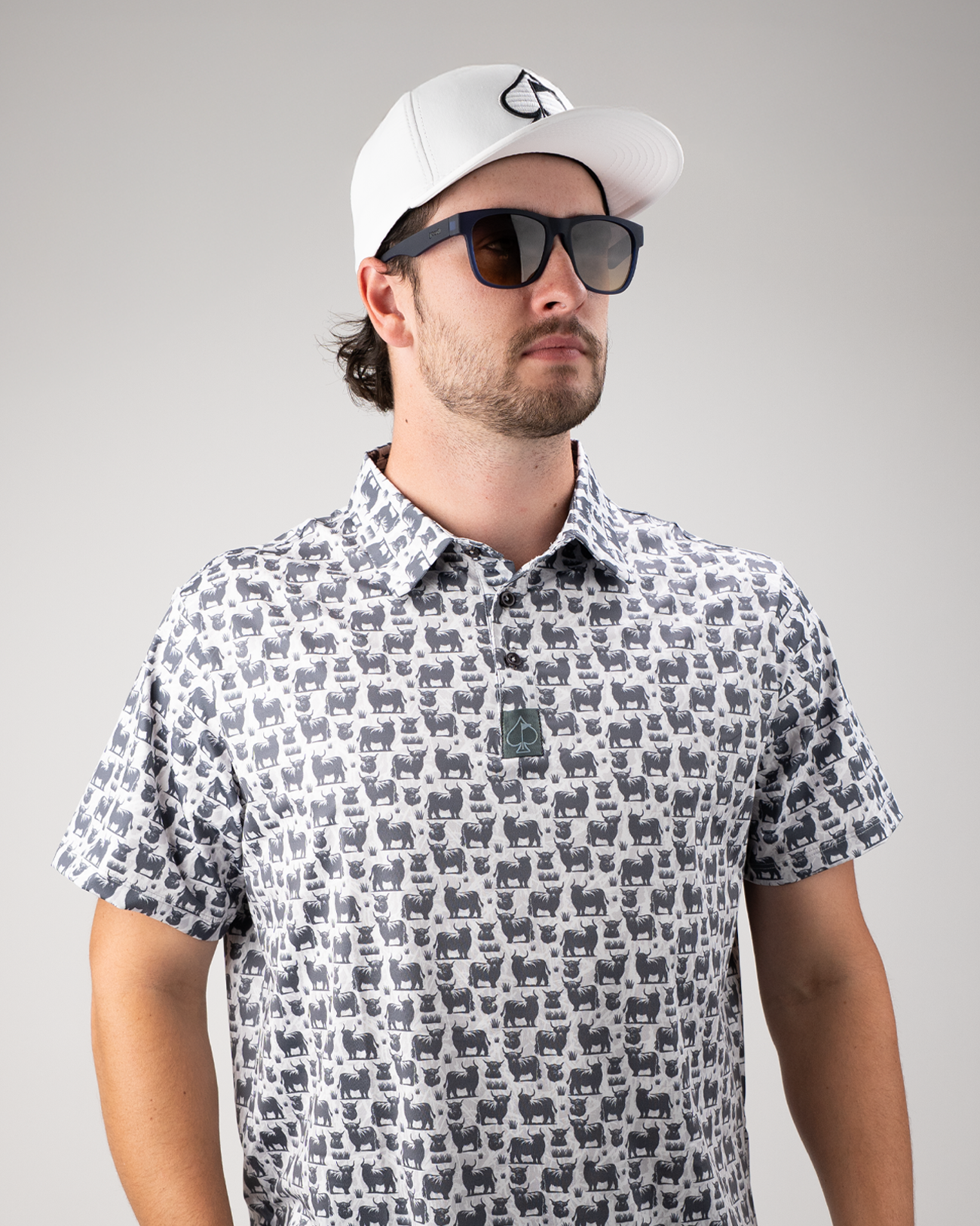 Man wearing a Highland Cow patterned golf shirt and a white cap, showcasing a stylish look with sunglasses.