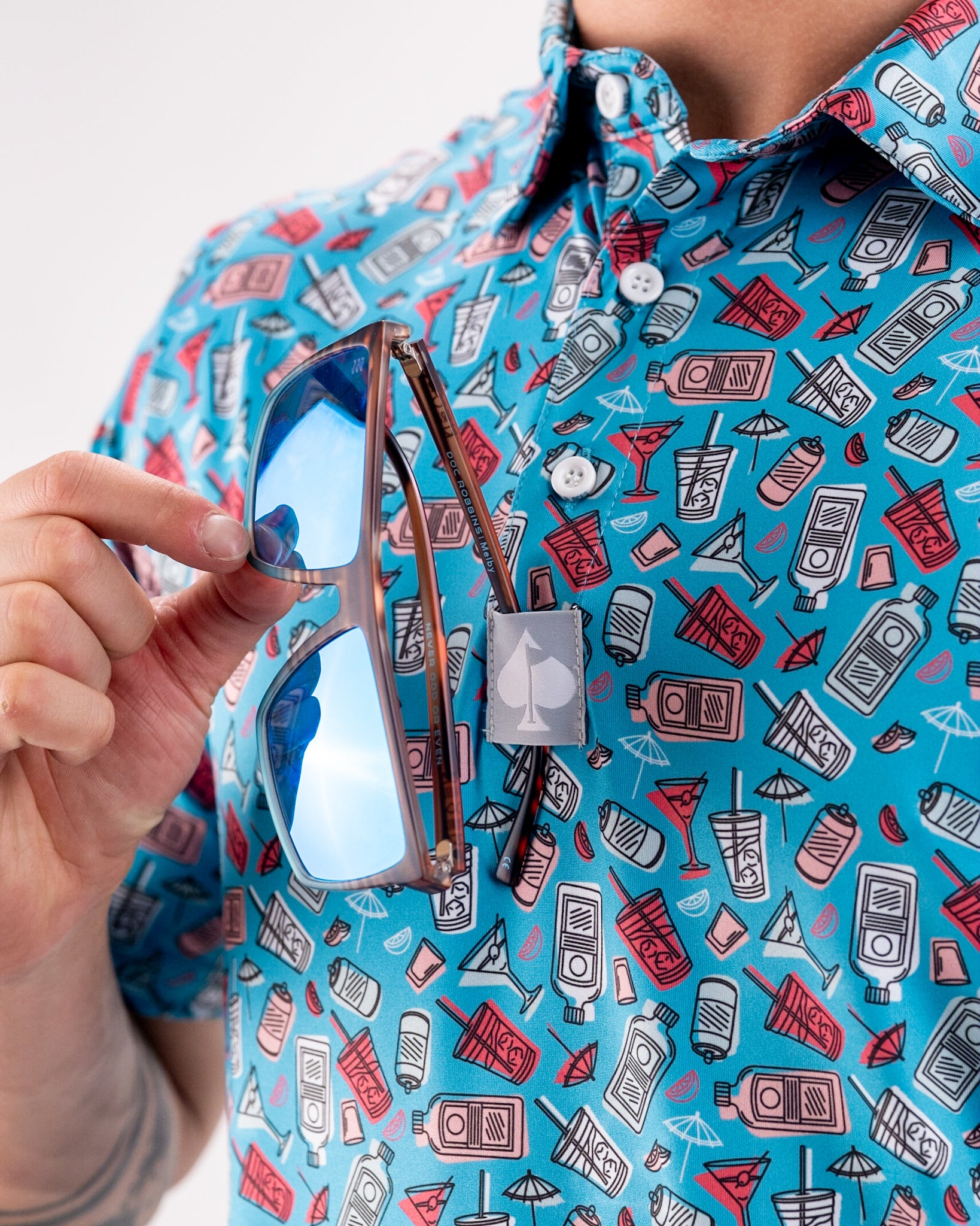 Bright blue golf shirt with drink and sun icon print, chest pocket with spade logo, holding mirrored sunglasses.