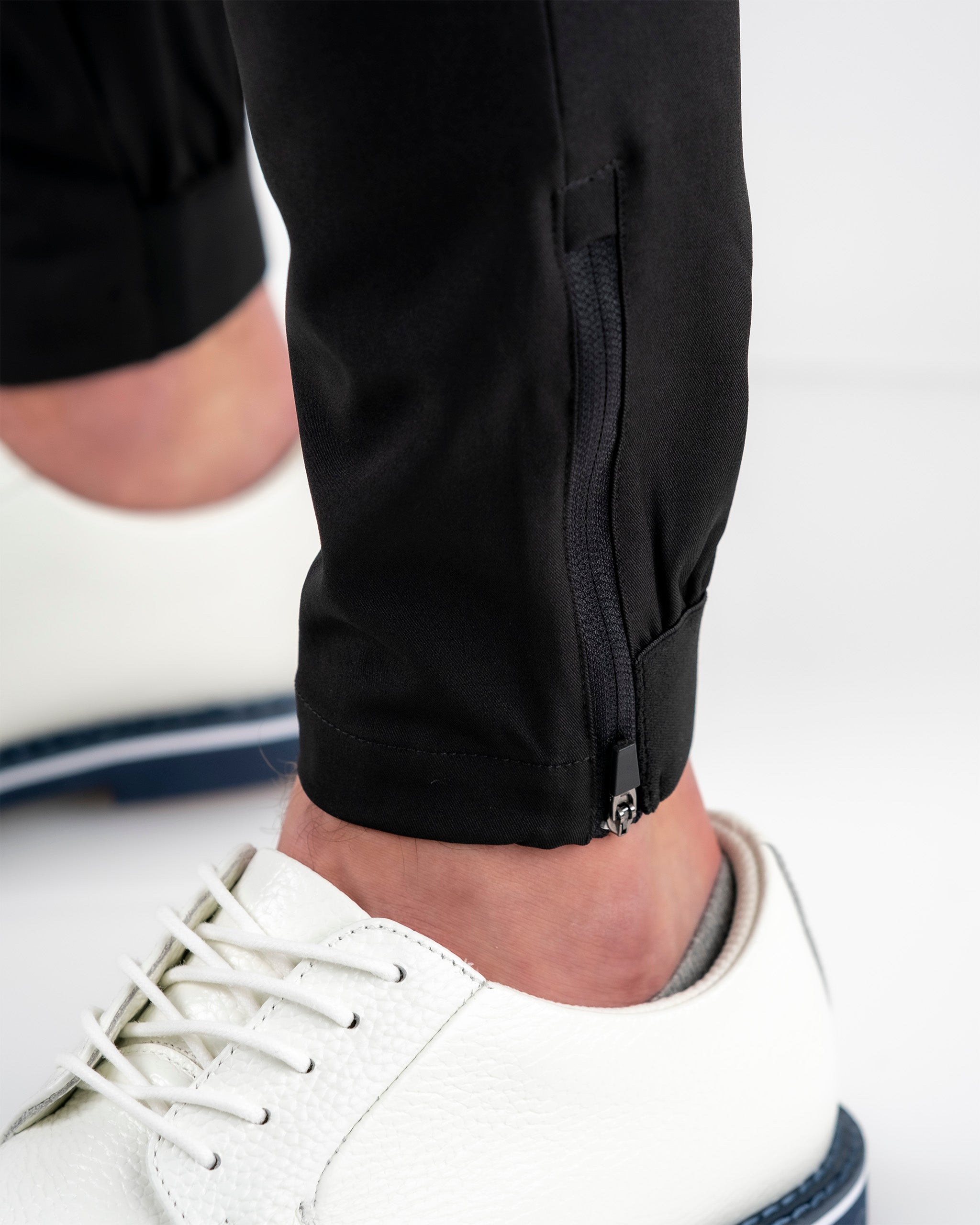 Close-up of a black golf pant with a zipper ankle detail, paired with white textured sneakers for a bold golf look.