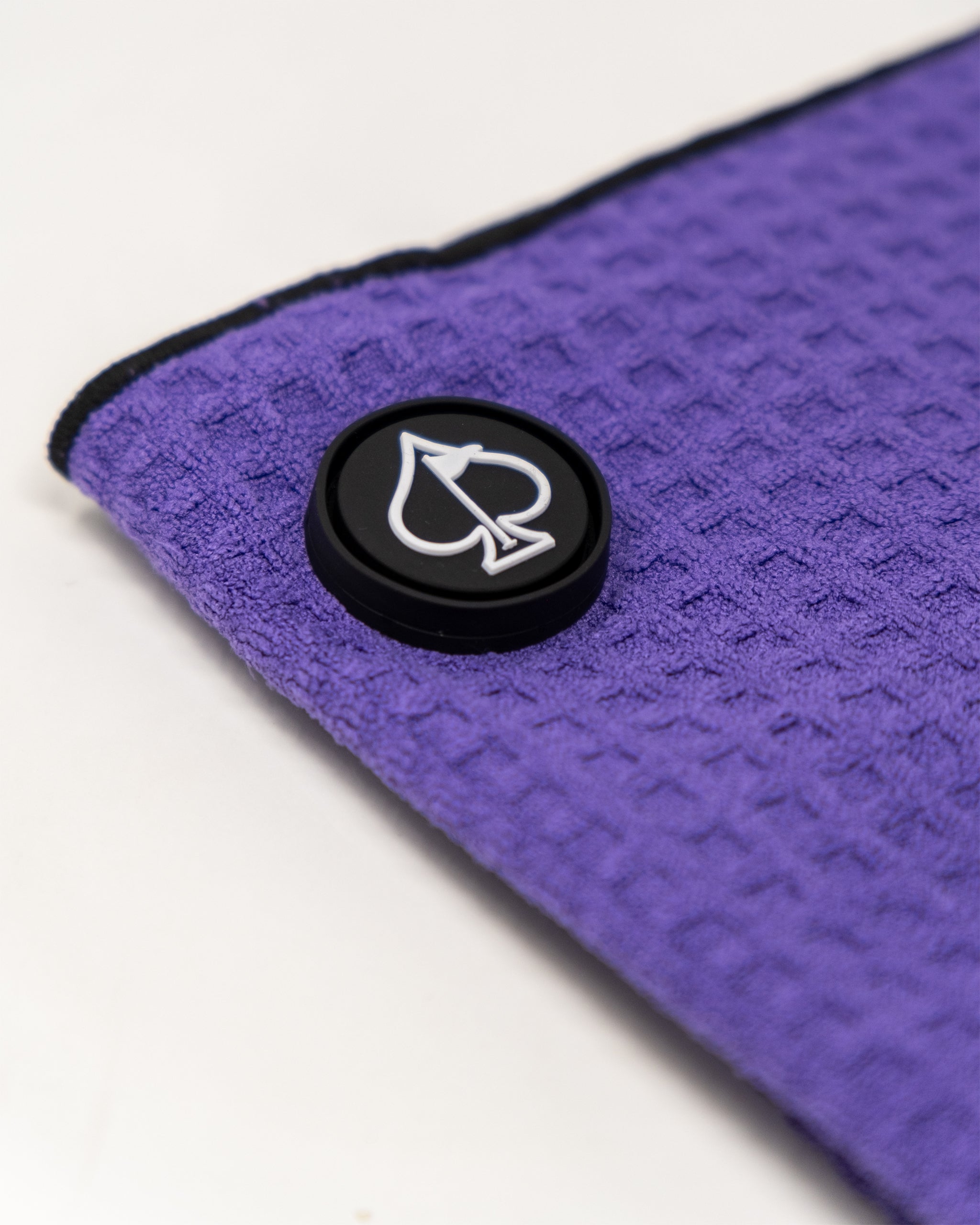 Close-up of a purple textured golf towel with a black button featuring a white spade and arrow logo, highlighting bold gol...