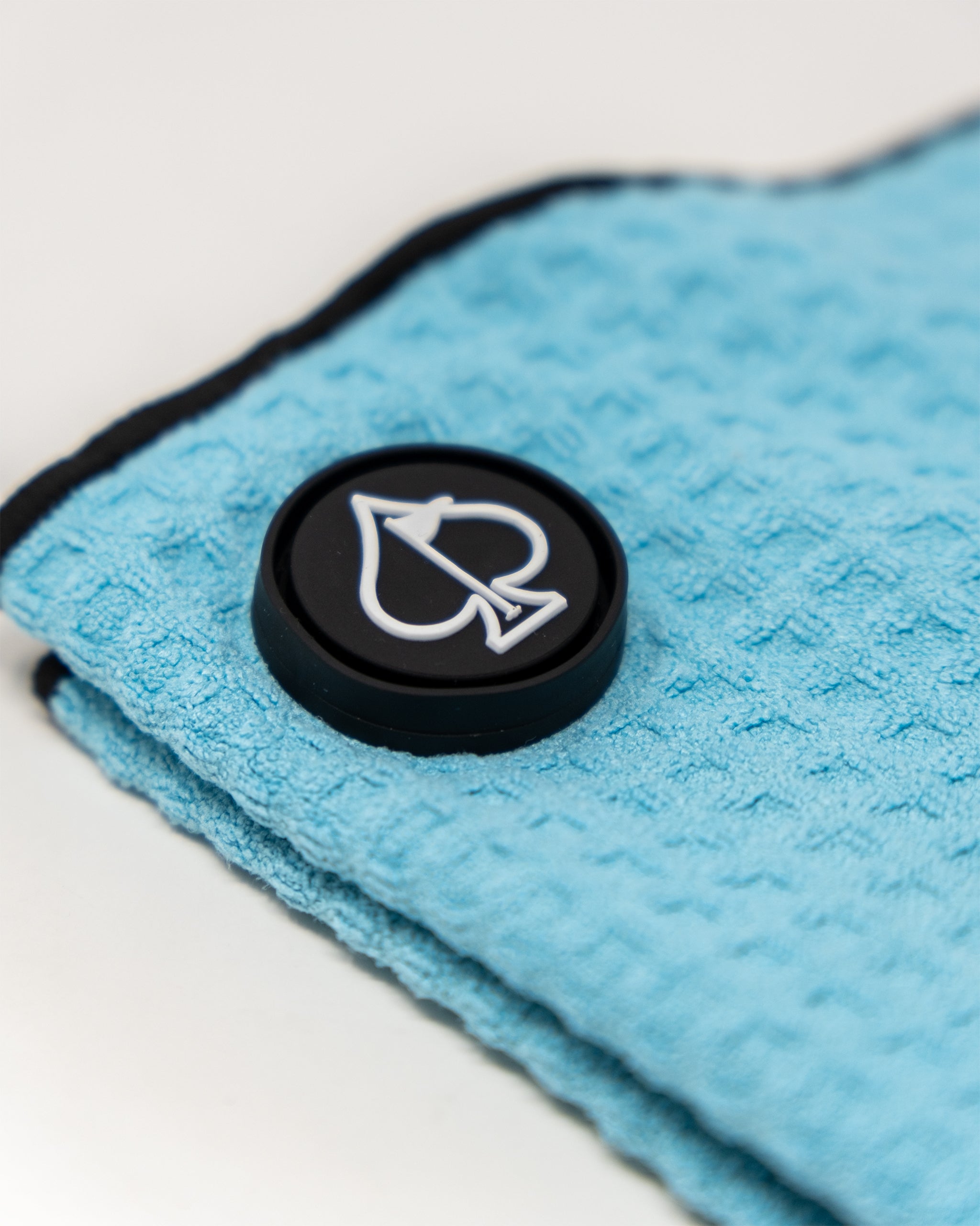 Close-up of a blue microfiber golf towel with a black logo button featuring a white arrow and shield design.
