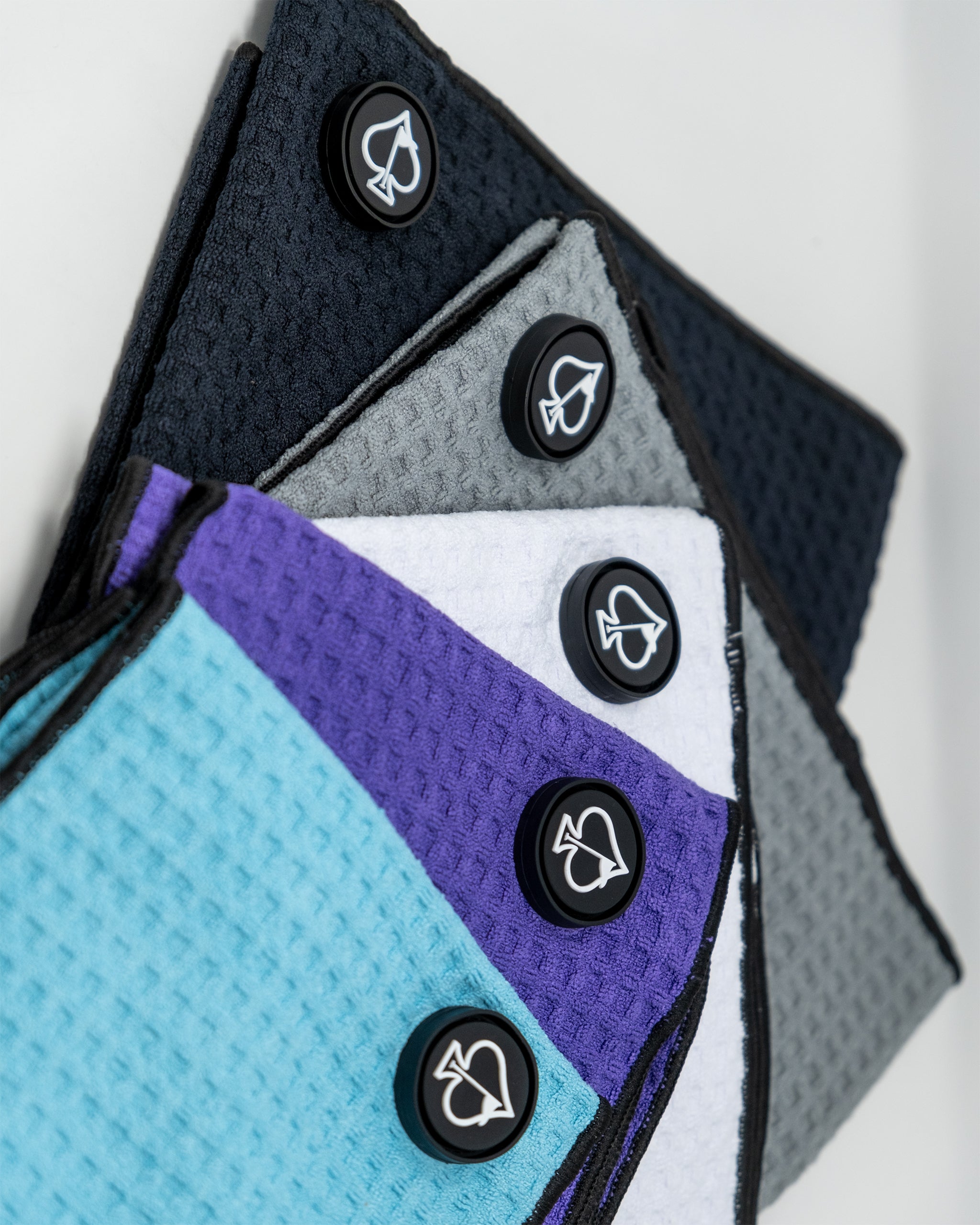 Colorful golf towels in black, gray, white, purple, and blue with bold spade logo buttons for stylish golf accessories.