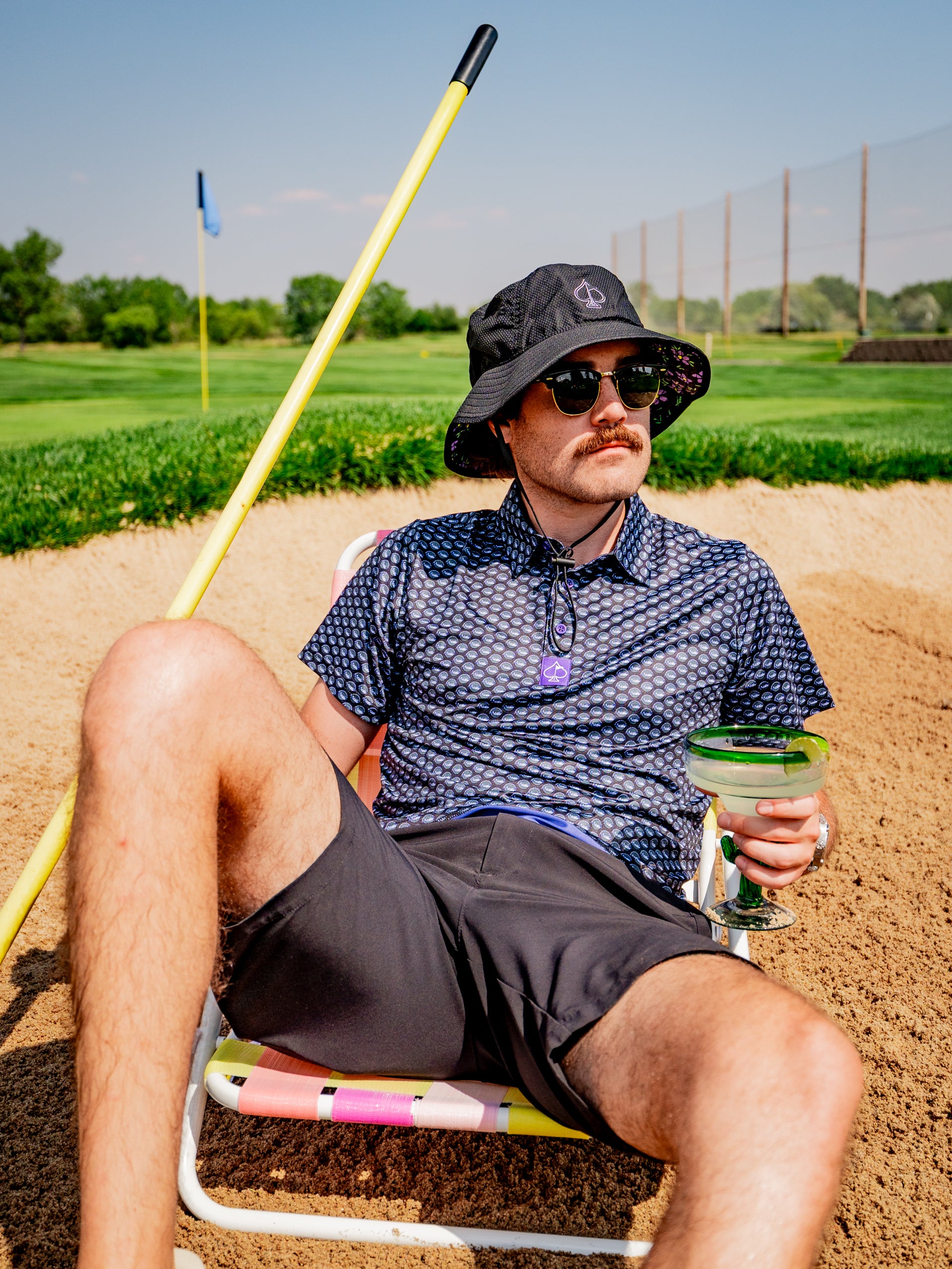 Man lounging in a golf sand trap, wearing a patterned shirt and bucket hat, holding a margarita, exuding a relaxed vibe.