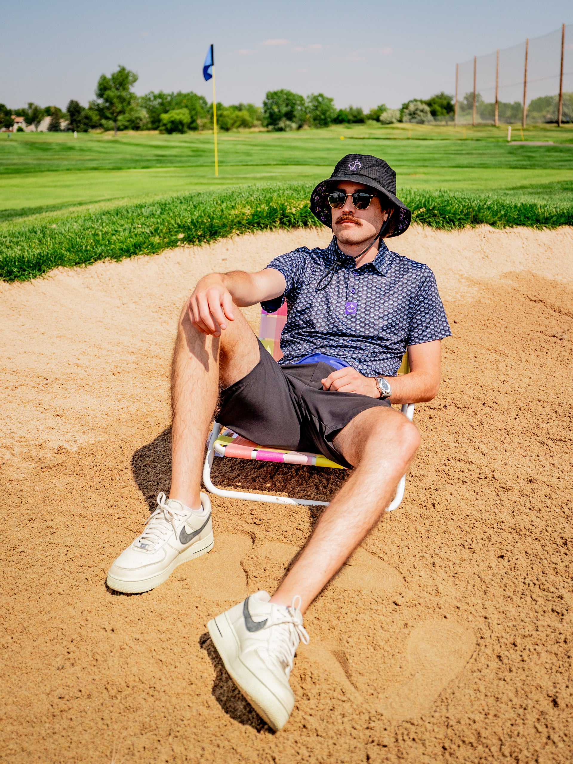 A golfer relaxes in a sand trap wearing a Margaritaville Hemisphere Dancer shirt, shorts, and a bucket hat, enjoying the sunny day.