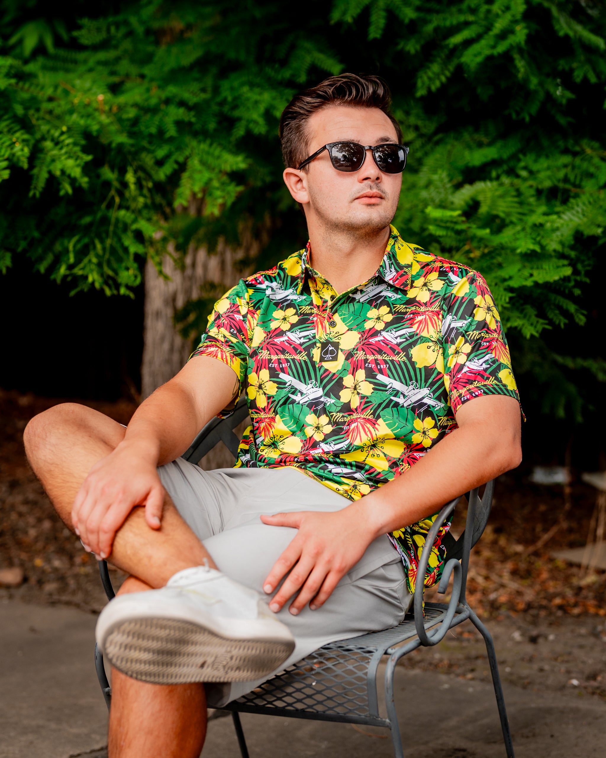 Man wearing a vibrant floral Margaritaville shirt and sunglasses, sitting casually in a chair surrounded by greenery.