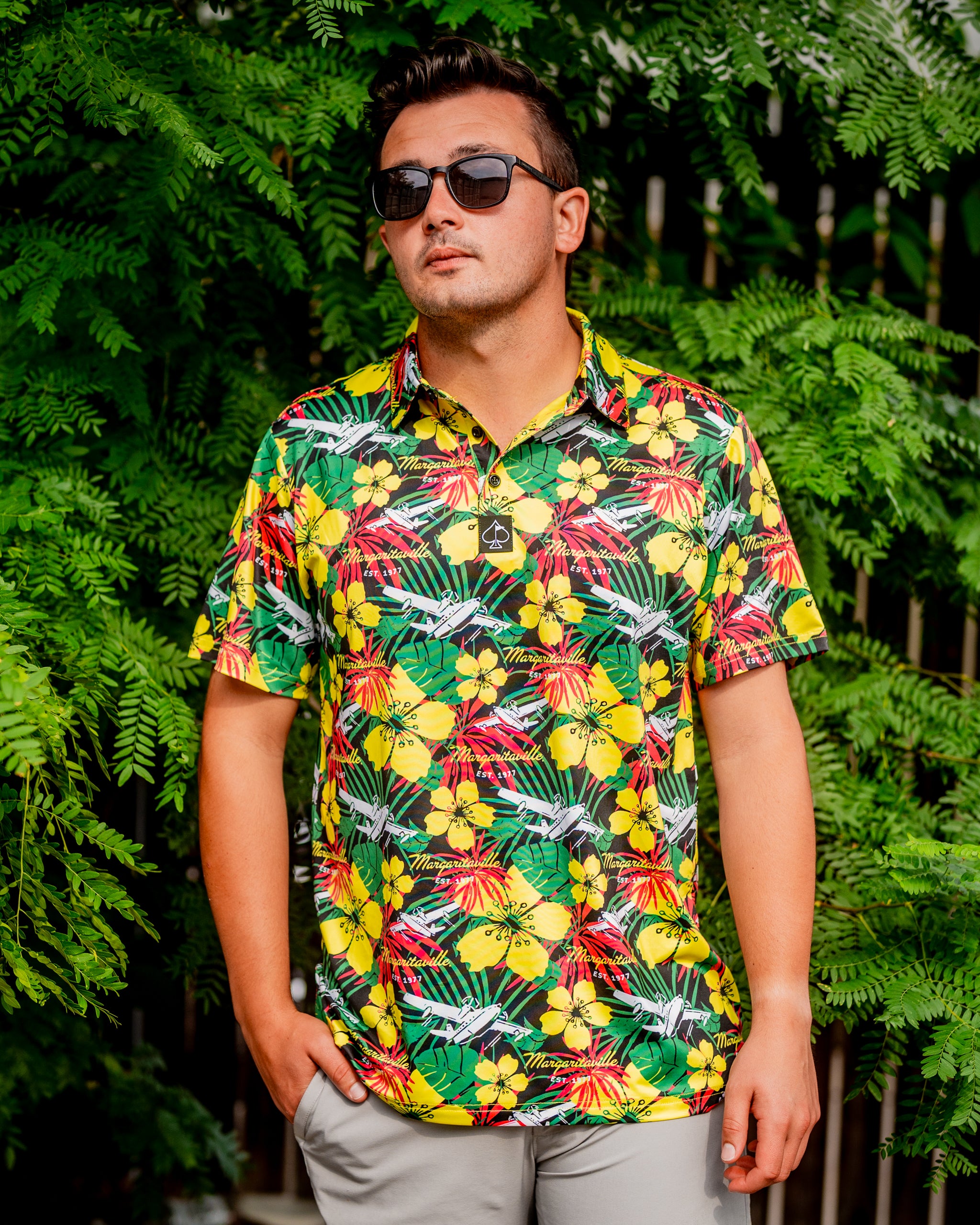 A man wearing a vibrant, floral-patterned polo shirt stands confidently against a lush green backdrop.