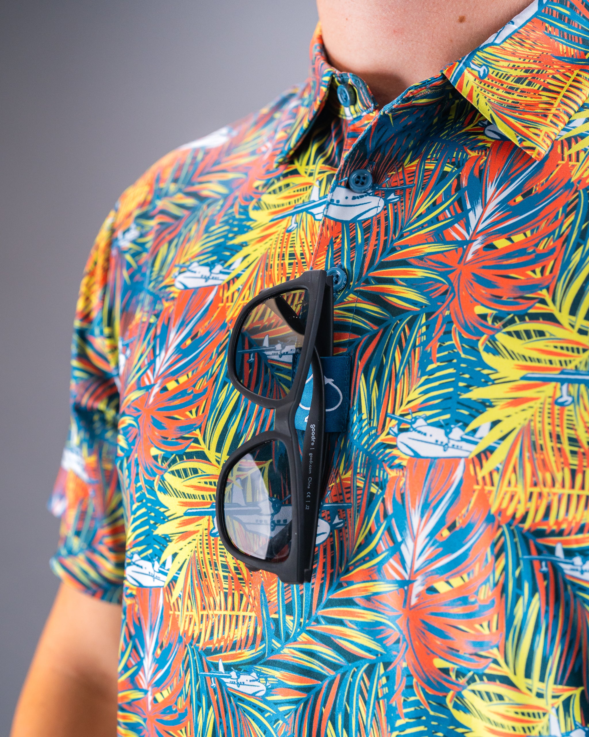 Bright, tropical print golf shirt with colorful palm leaves and small boat illustrations, paired with black sunglasses.