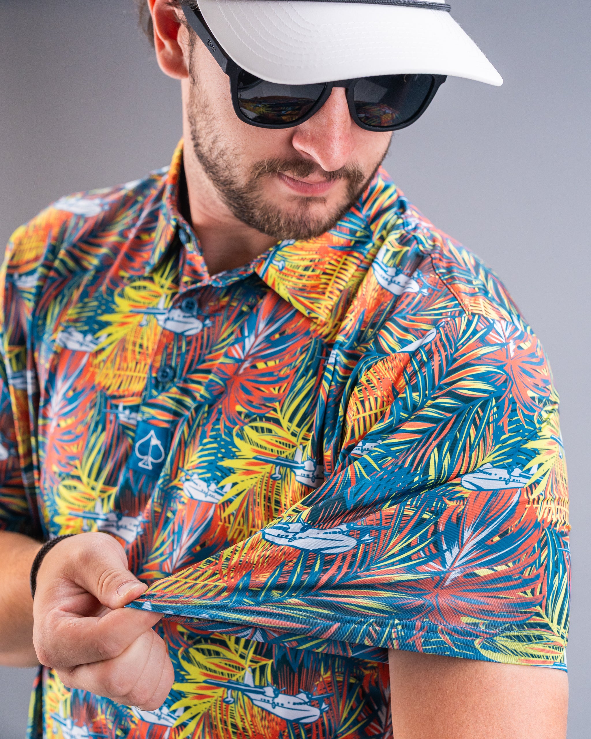 Man in colorful, tropical-themed golf shirt with palm leaves and boats, wearing sunglasses and white cap, showcasing bold ...