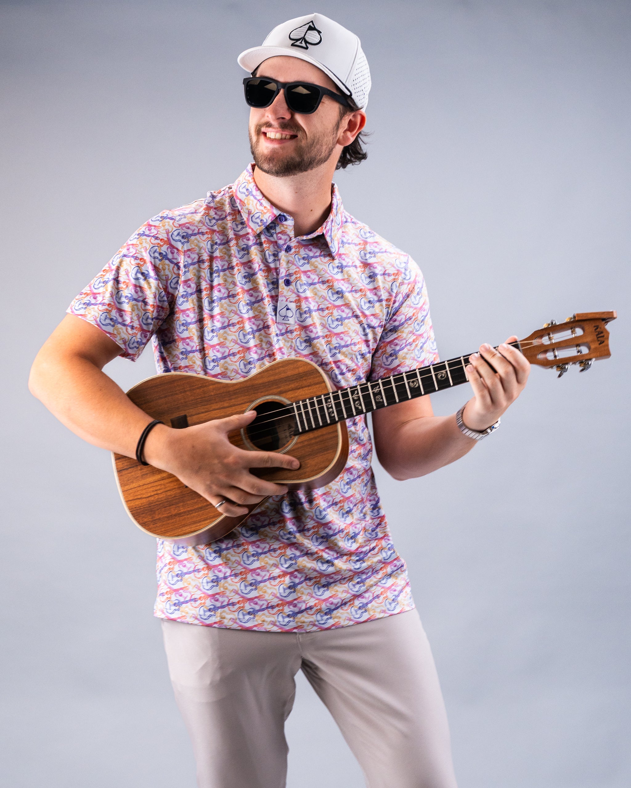 Man wearing a colorful, patterned golf shirt, sunglasses, and a white cap with a spade logo, playing a wooden ukulele.