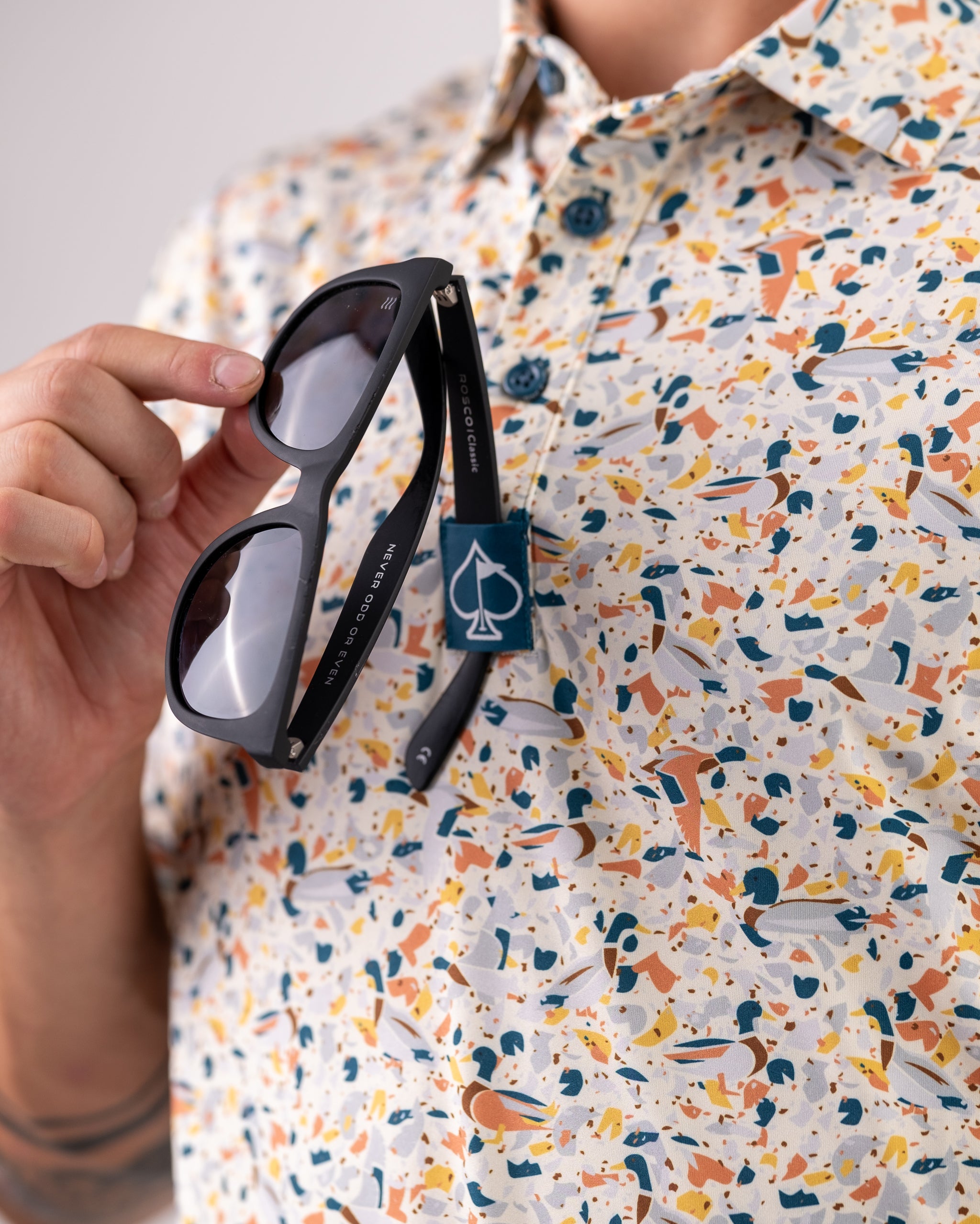 Close-up of a person holding black sunglasses with "Never Do or Do Even" on the arm, wearing a colorful, bold patterned go...