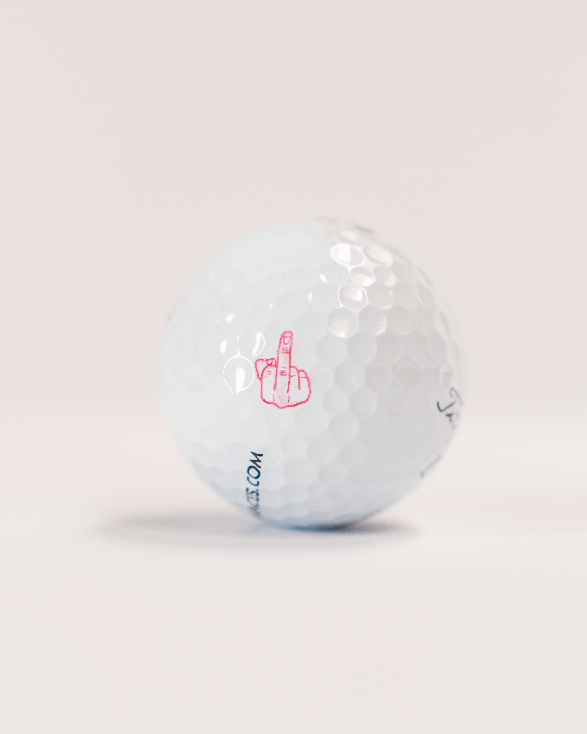 Golf ball with a red middle finger stamp graphic and partial website text, designed for humorous or novelty use.