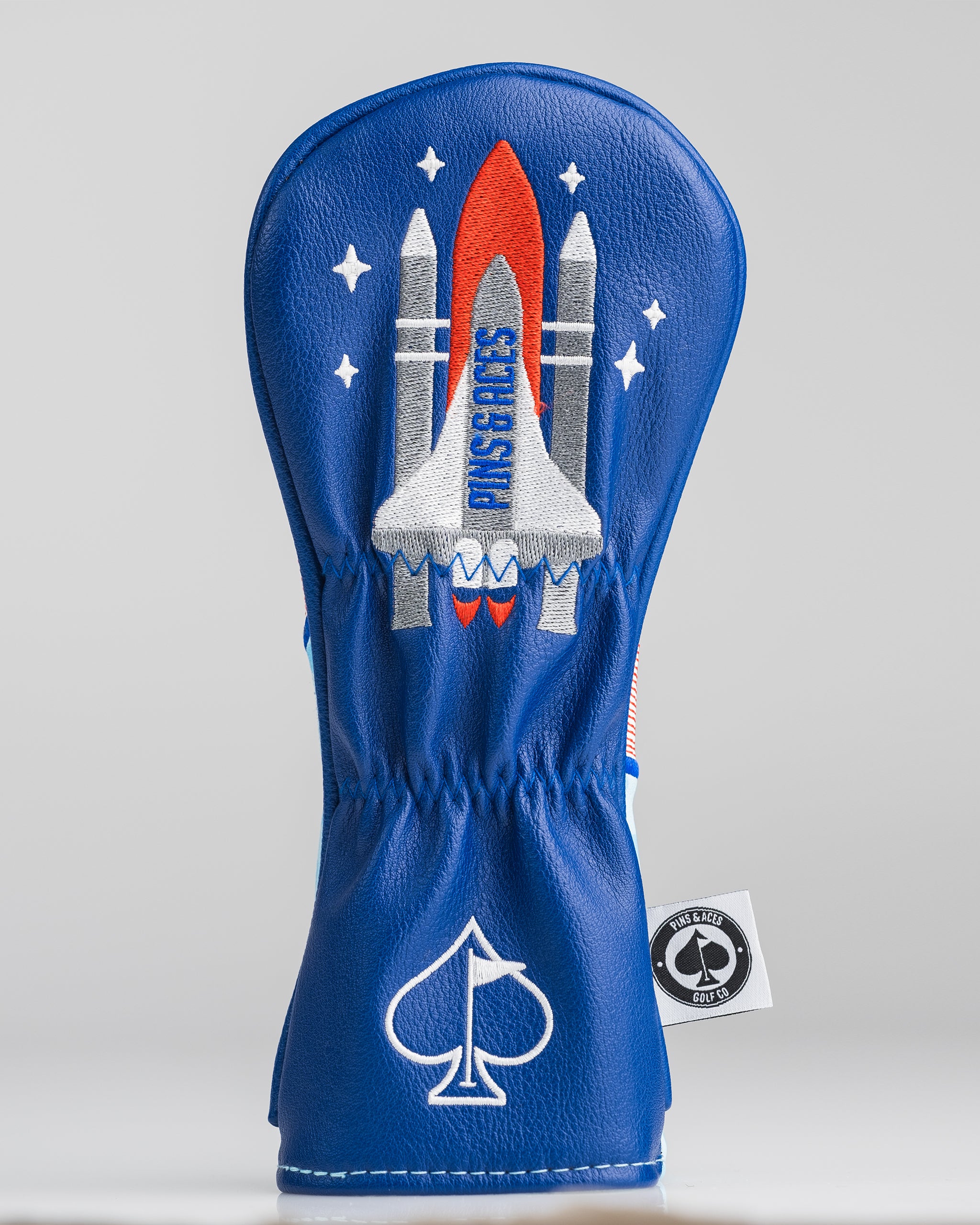 Blue golf headcover with embroidered rocket and "PINS & ACES" text, featuring stars and a spade logo, for bold golf style.