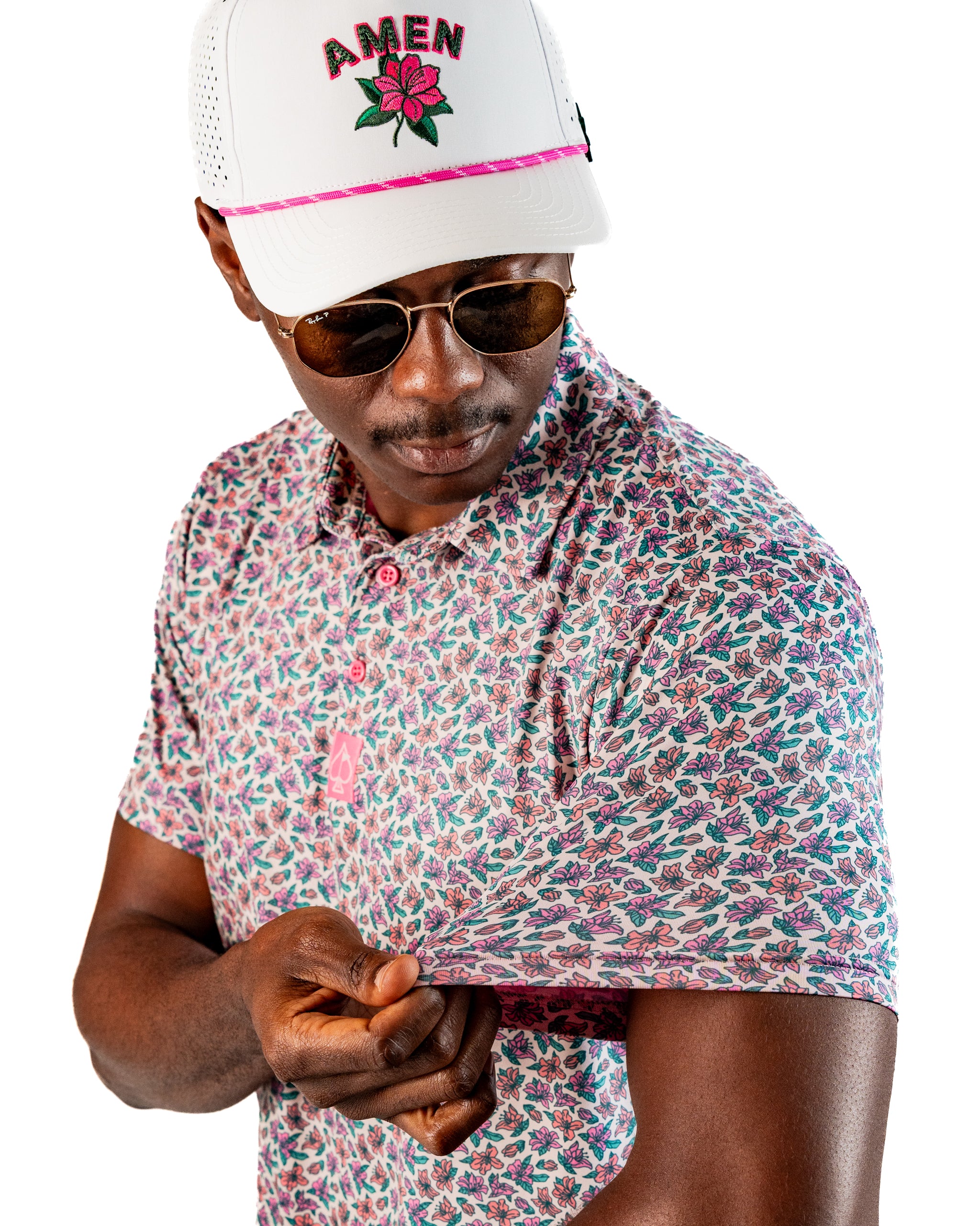 Man wearing a floral-patterned golf polo shirt in light azalea color, paired with sunglasses and a white cap.