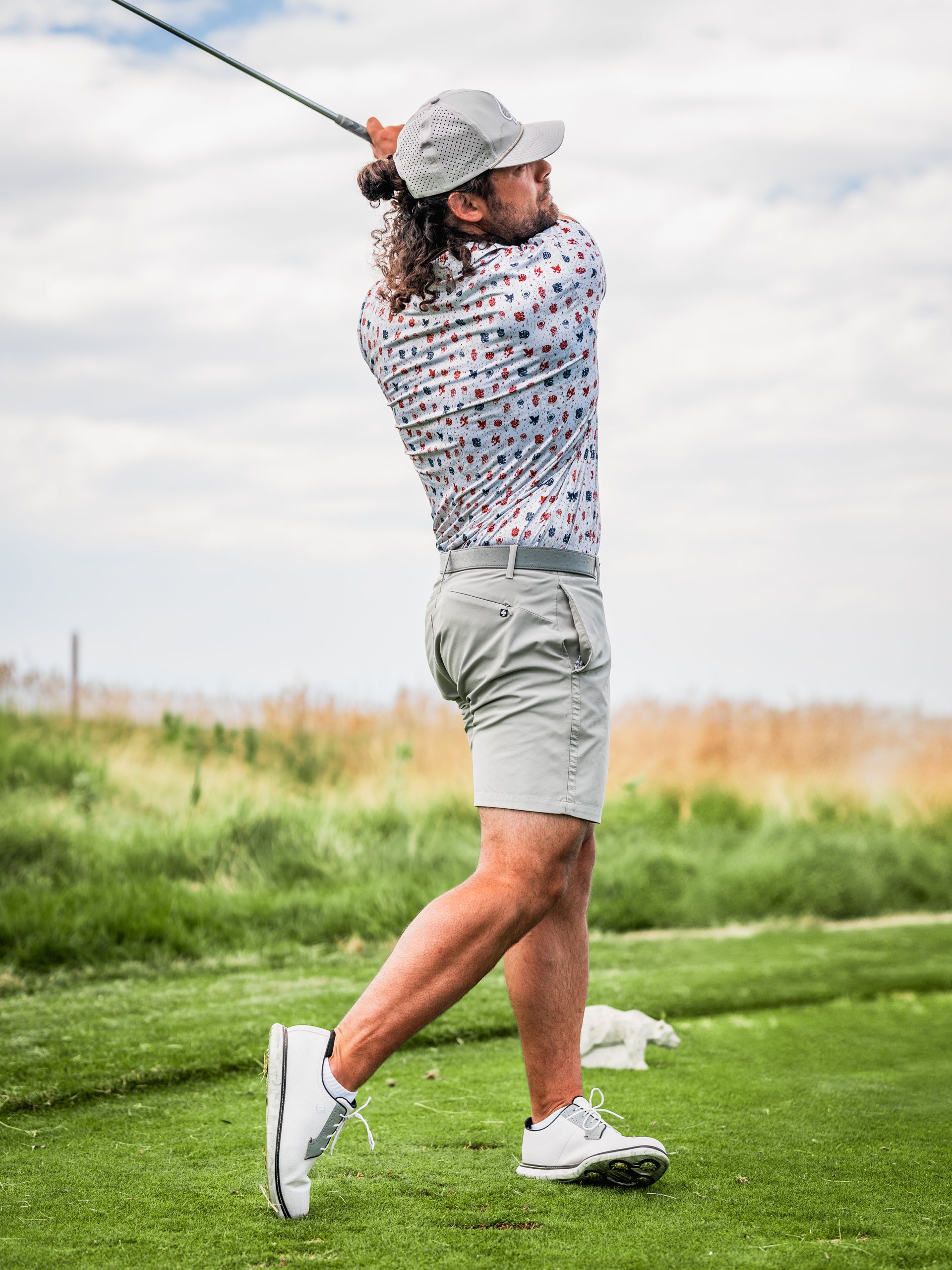 A golfer swings a club on the course, wearing a patterned shirt and light shorts, showcasing bold golf apparel.