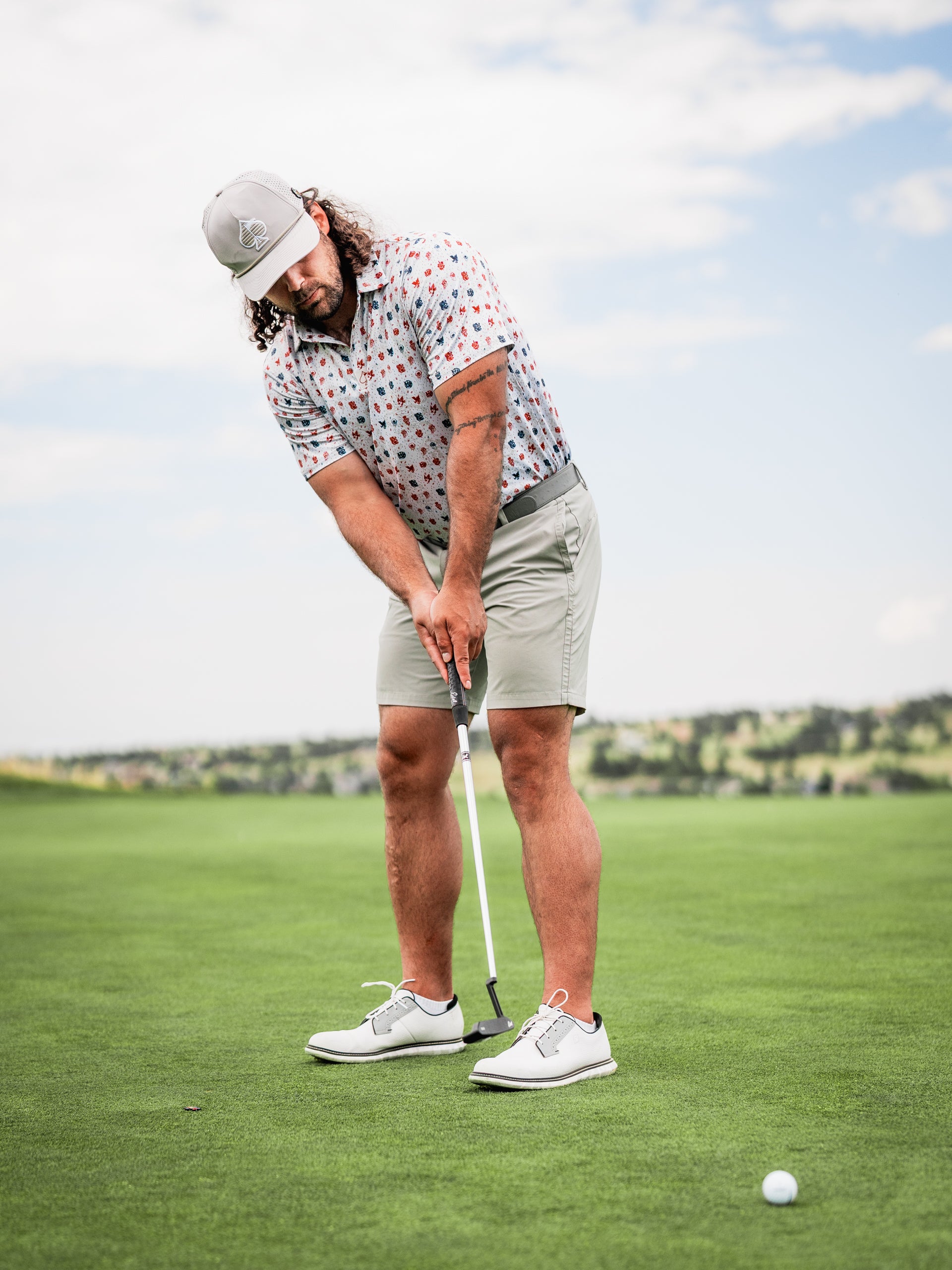 A golfer in a colorful patterned shirt and shorts prepares to putt on a lush green course under a blue sky.
