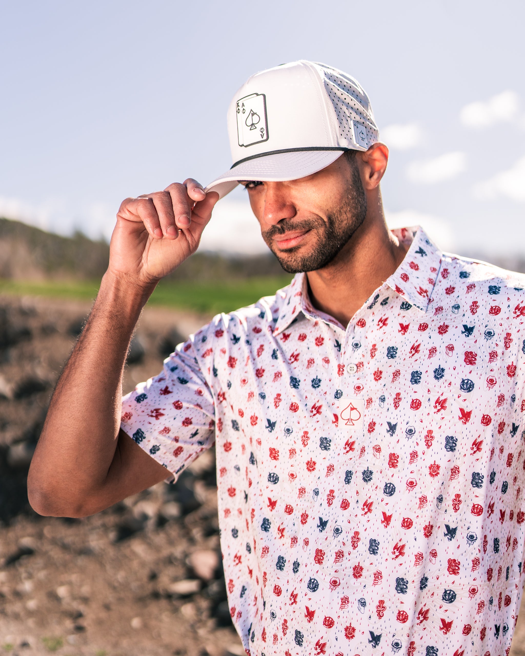 Man wearing a short-sleeve American Traditional golf shirt with a playful design and a white cap, adjusting his hat outdoors.