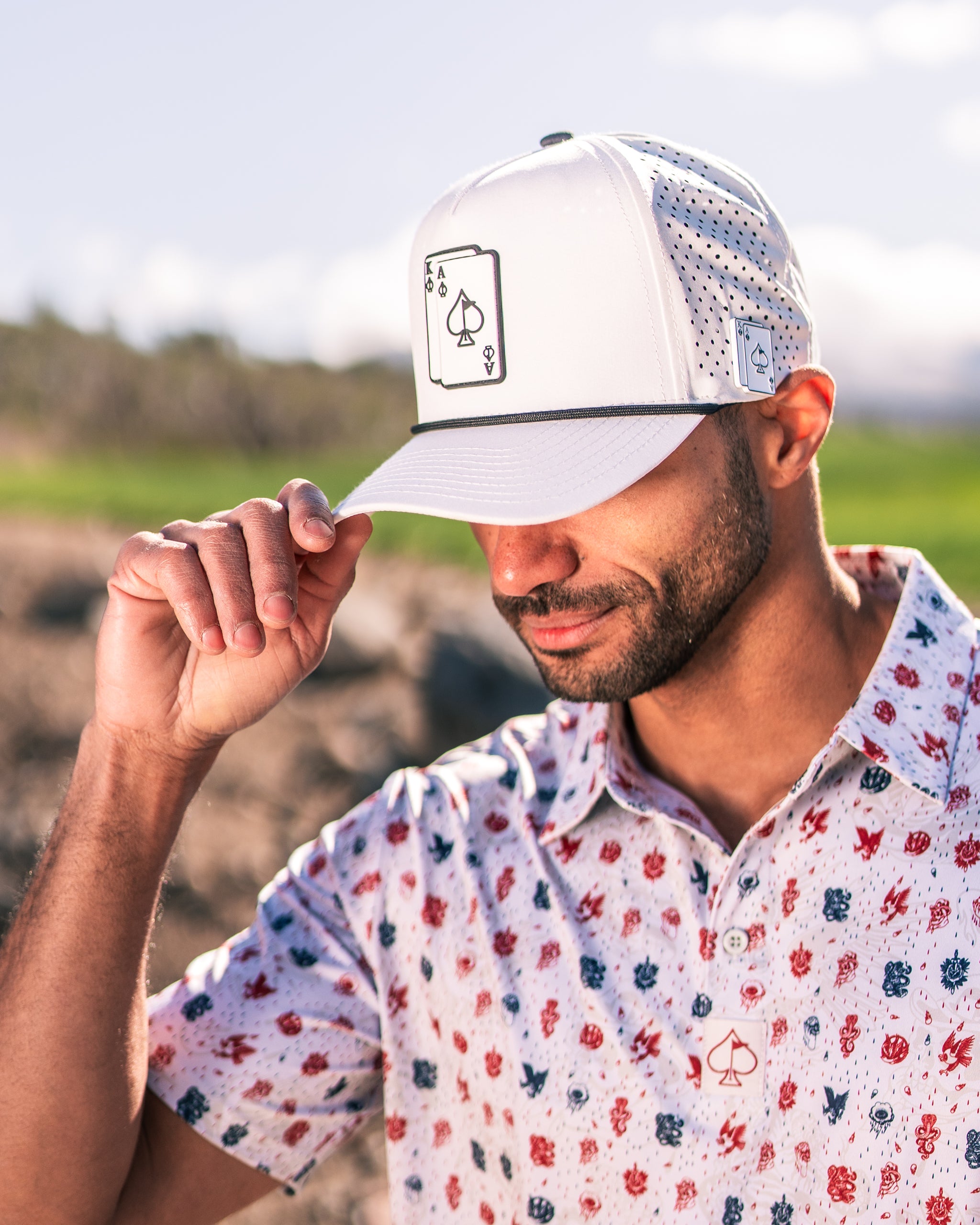Man adjusting a white mesh cap with an ace of spades logo, wearing a colorful floral golf shirt on a sunny day.
