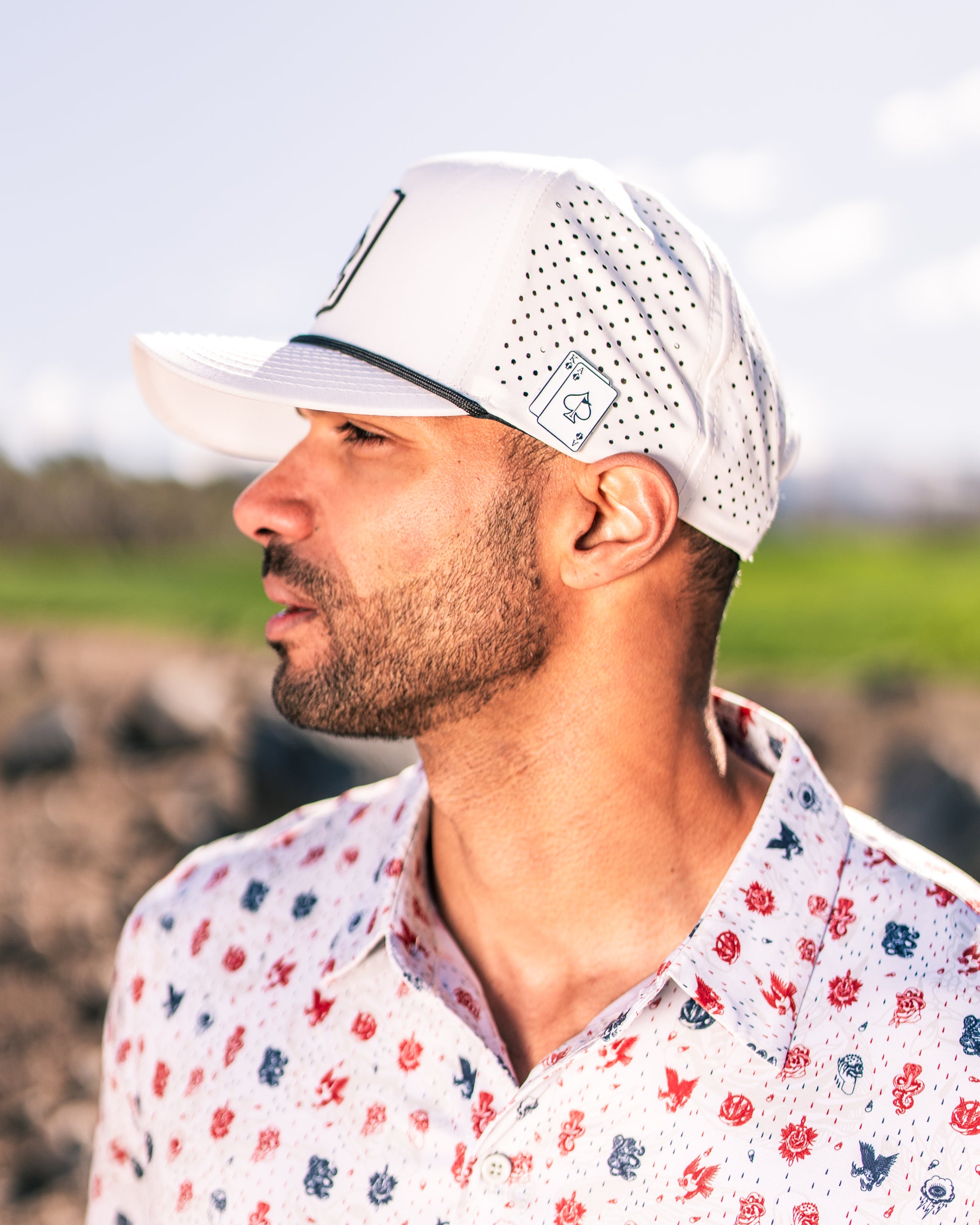 Man wearing a white golf cap with perforated design and a floral button-up shirt, set against a green landscape.