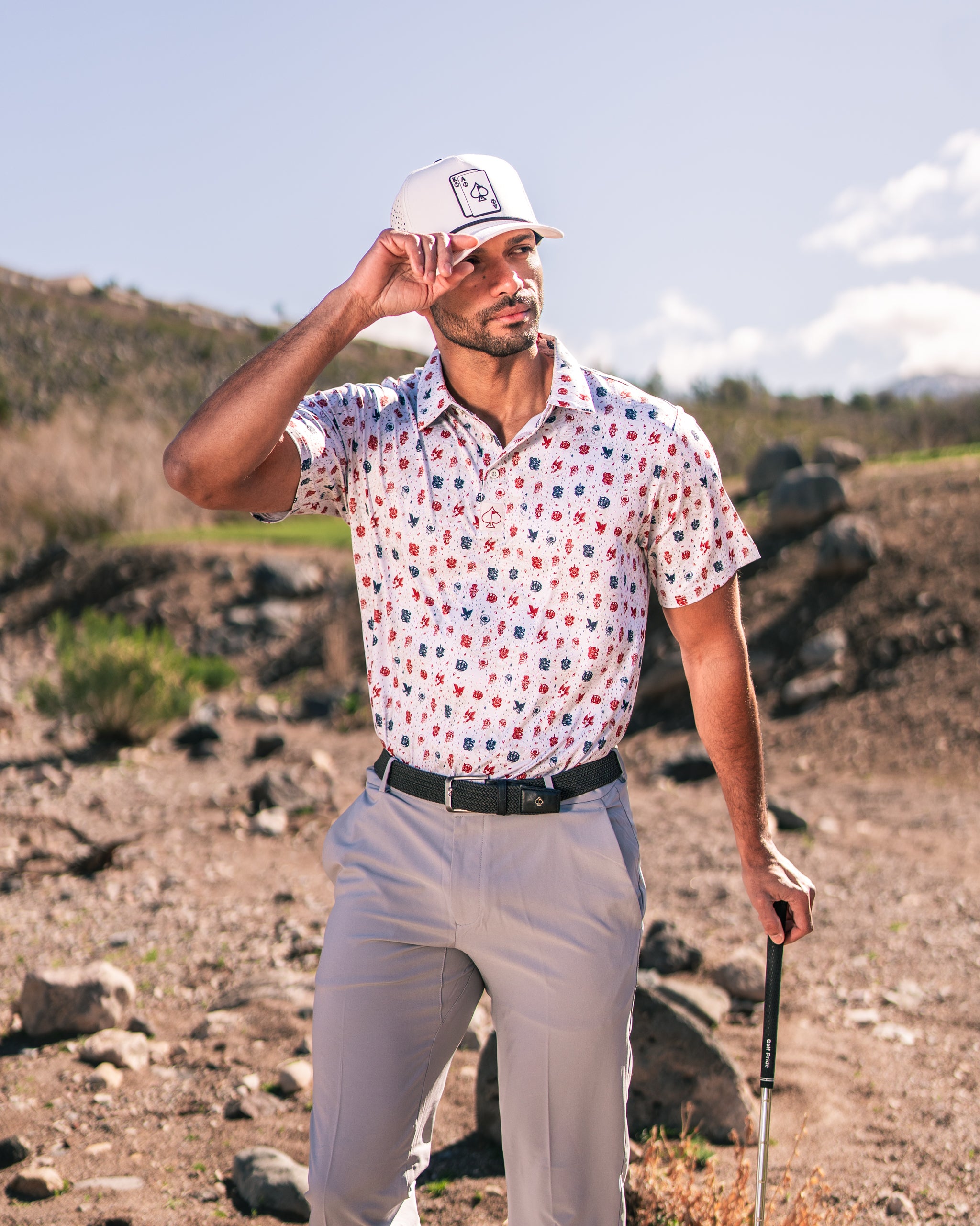 A golfer poses outdoors in a colorful American Traditional polo shirt, paired with light gray pants and a white cap.