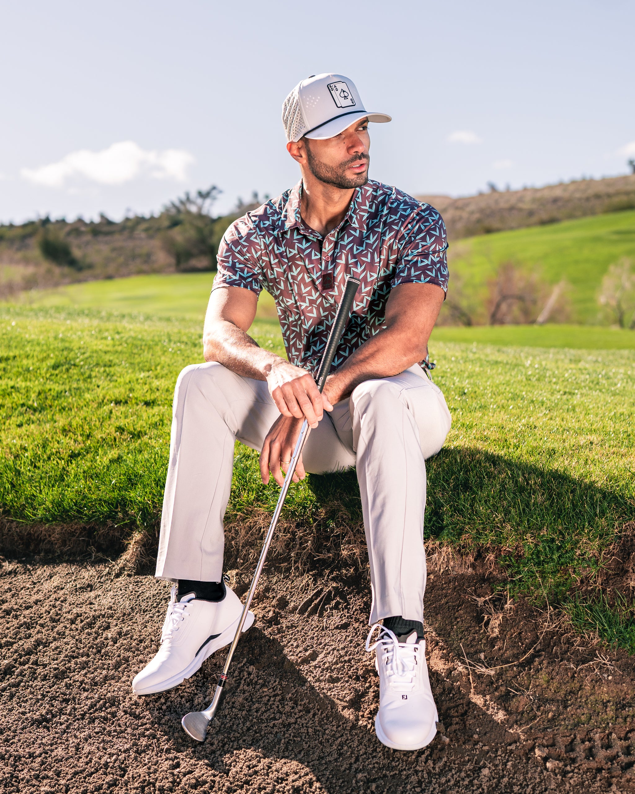 Man sitting on the grass in a colorful golf shirt and light pants, holding a golf club, with a scenic course backdrop.