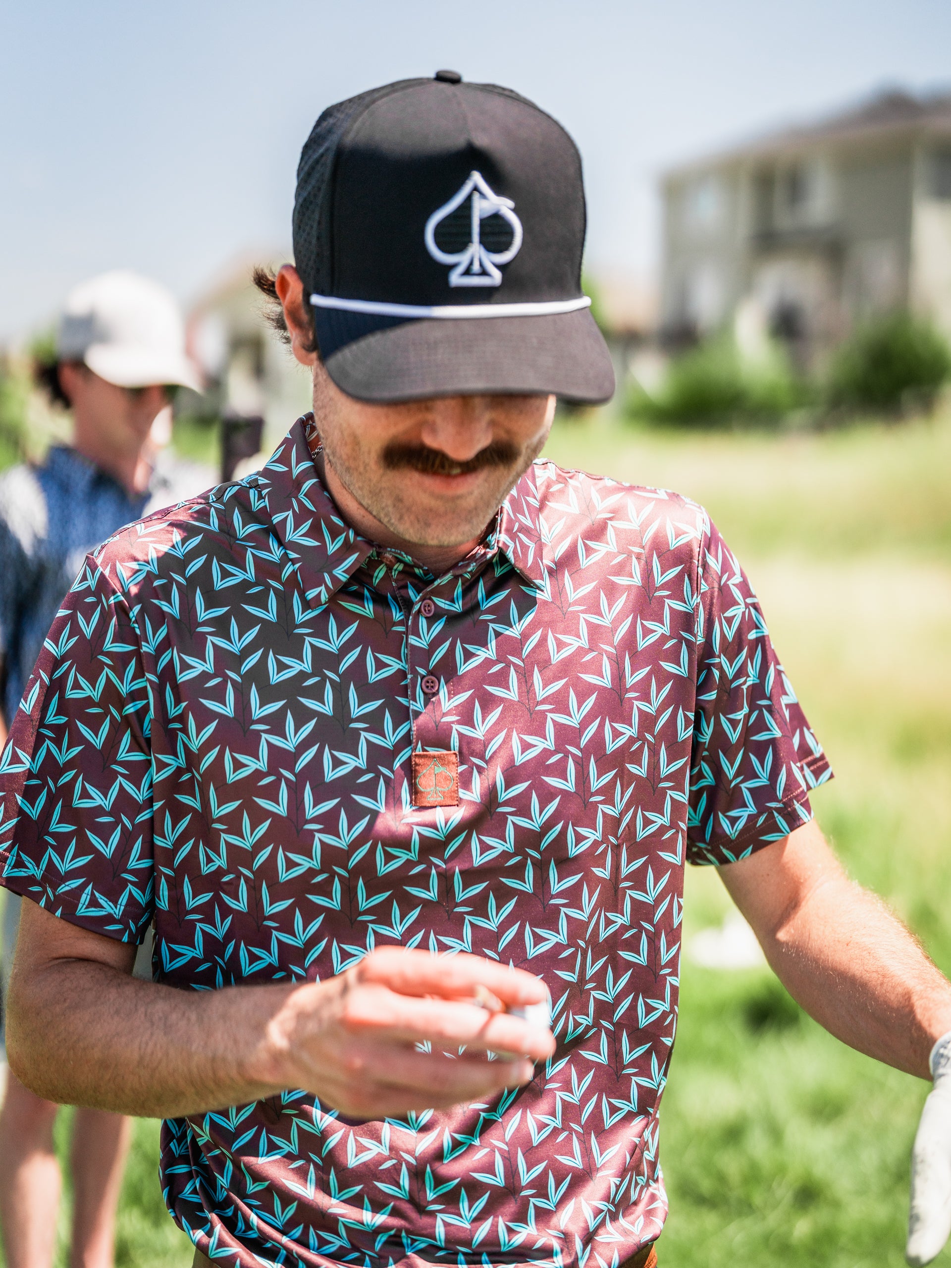 A golfer wearing a patterned Cocoa Verde polo shirt and a black cap smiles while holding a drink on the course.