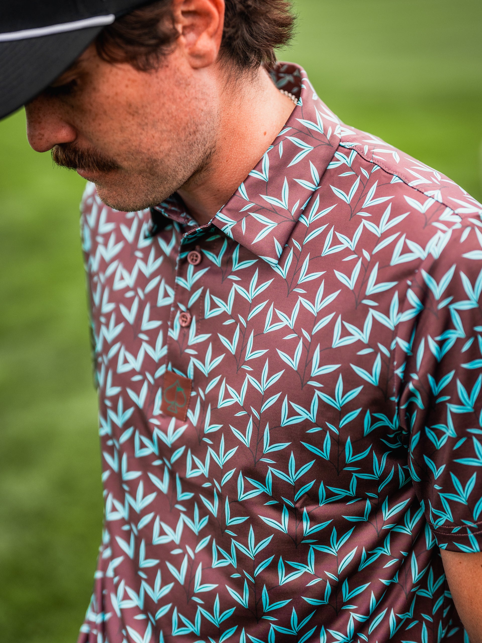 Men's Cocoa Verde golf polo featuring a bold brown and turquoise leaf pattern, perfect for stylish performance on the course.