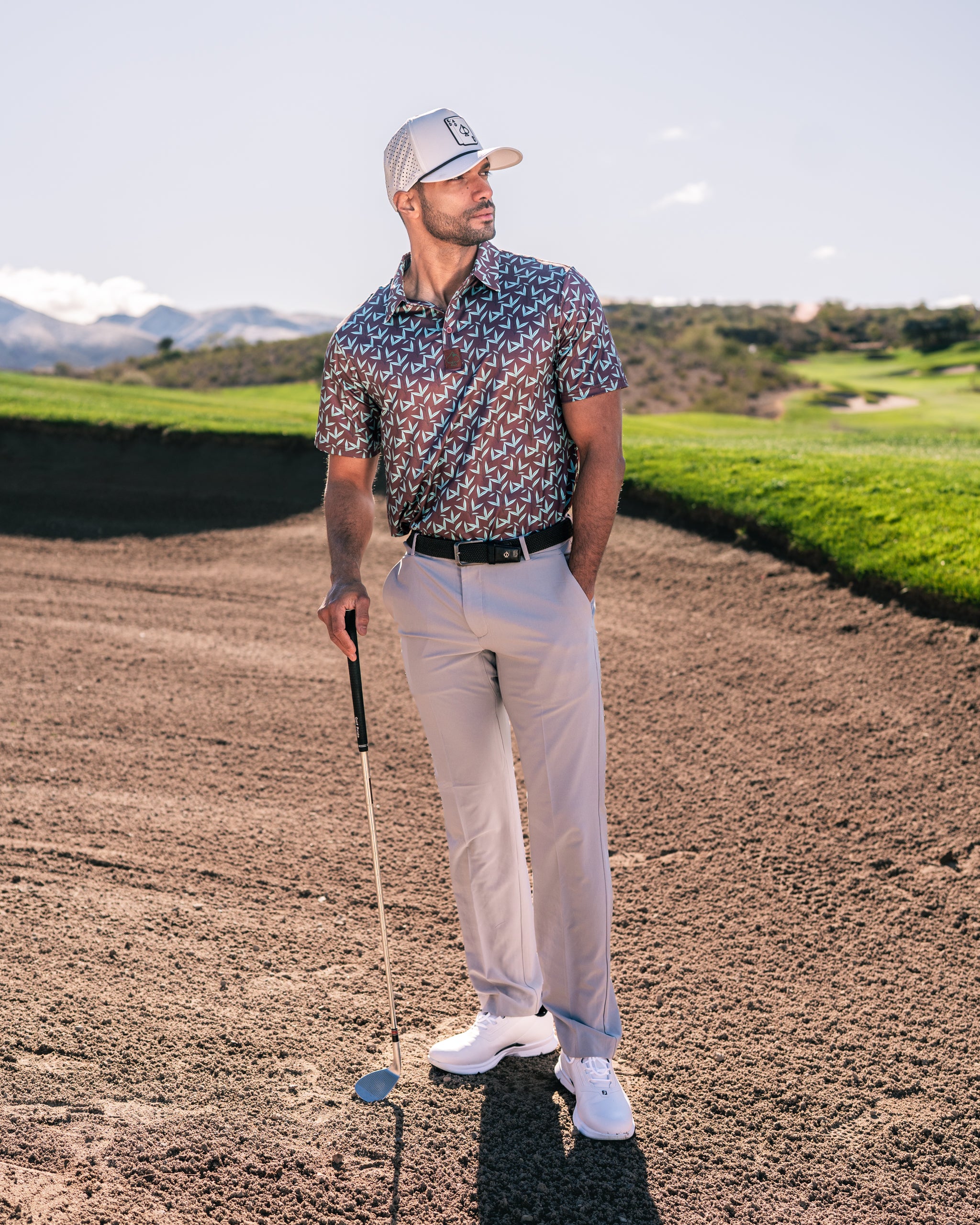Stylish golfer in Cocoa Verde polo and light pants stands on the course, ready to play with a club in hand.