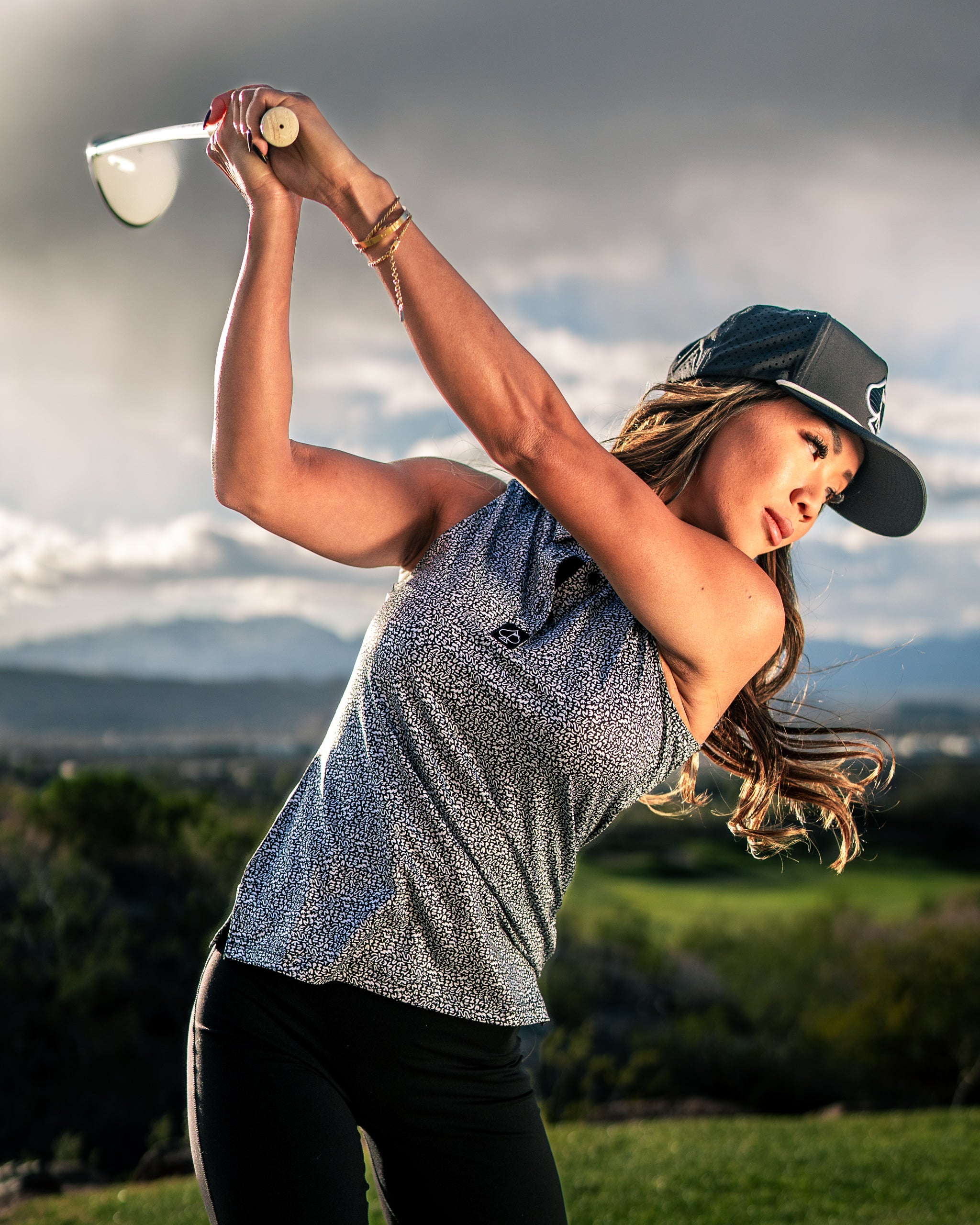 Woman in a stylish coral golf top and cap swings a driver on a scenic golf course, showcasing bold golf fashion.