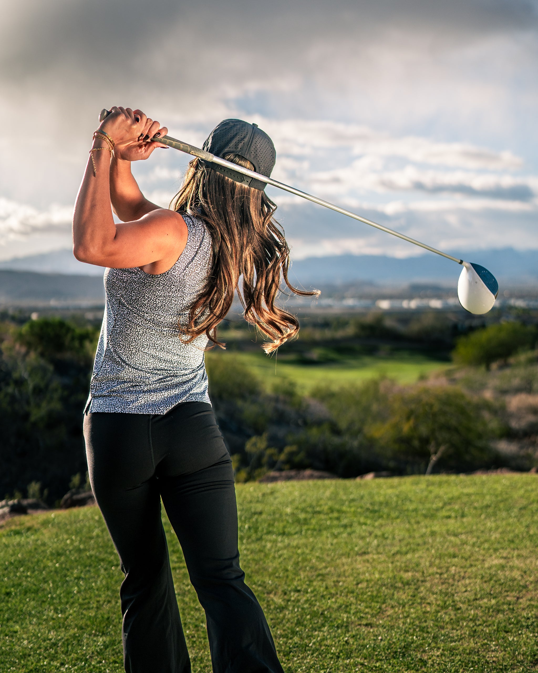Woman wearing a stylish coral golf top and black pants swings a driver on a scenic golf course.