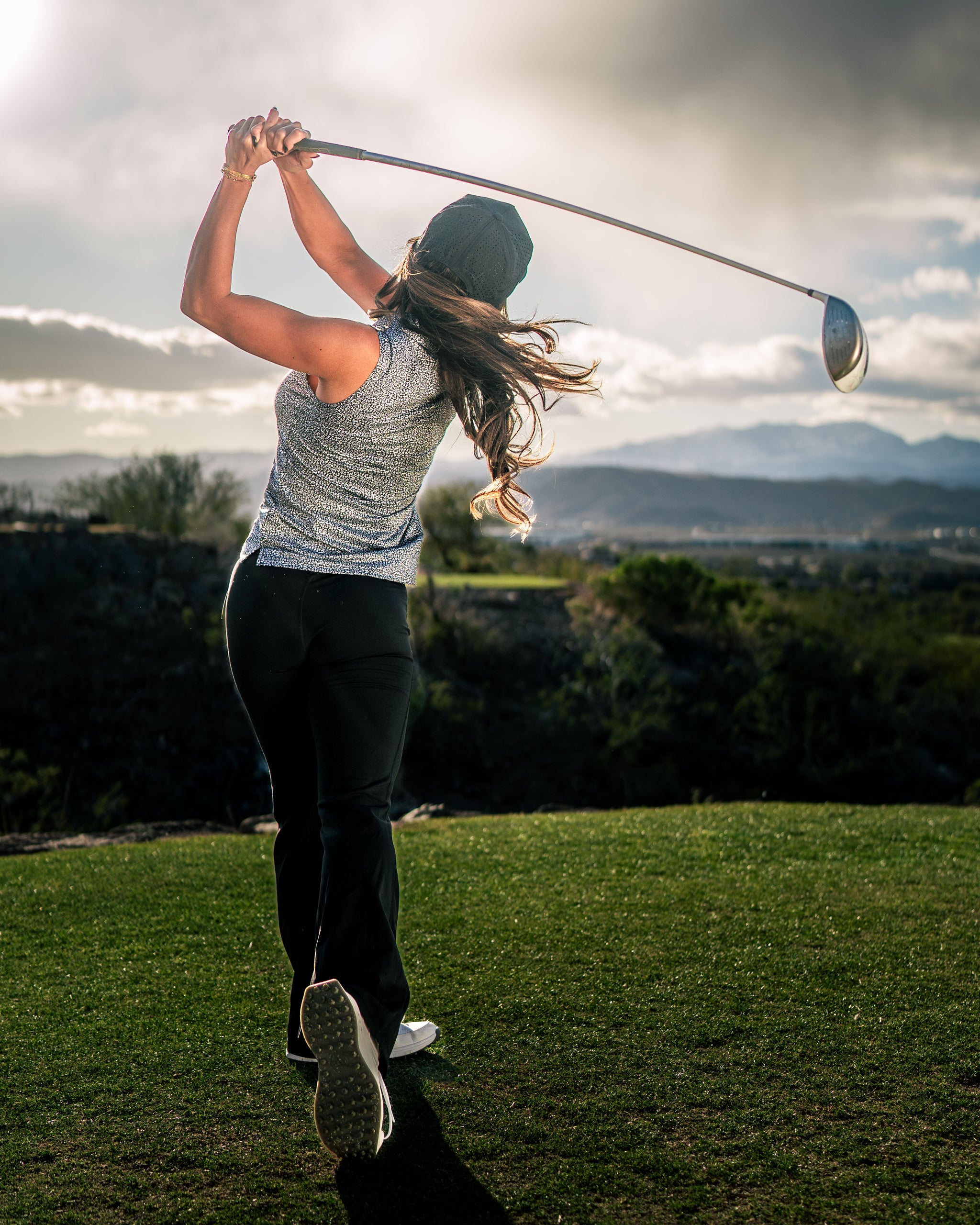 A woman swings a golf club on a course, wearing a stylish coral top and black pants, with a scenic backdrop.