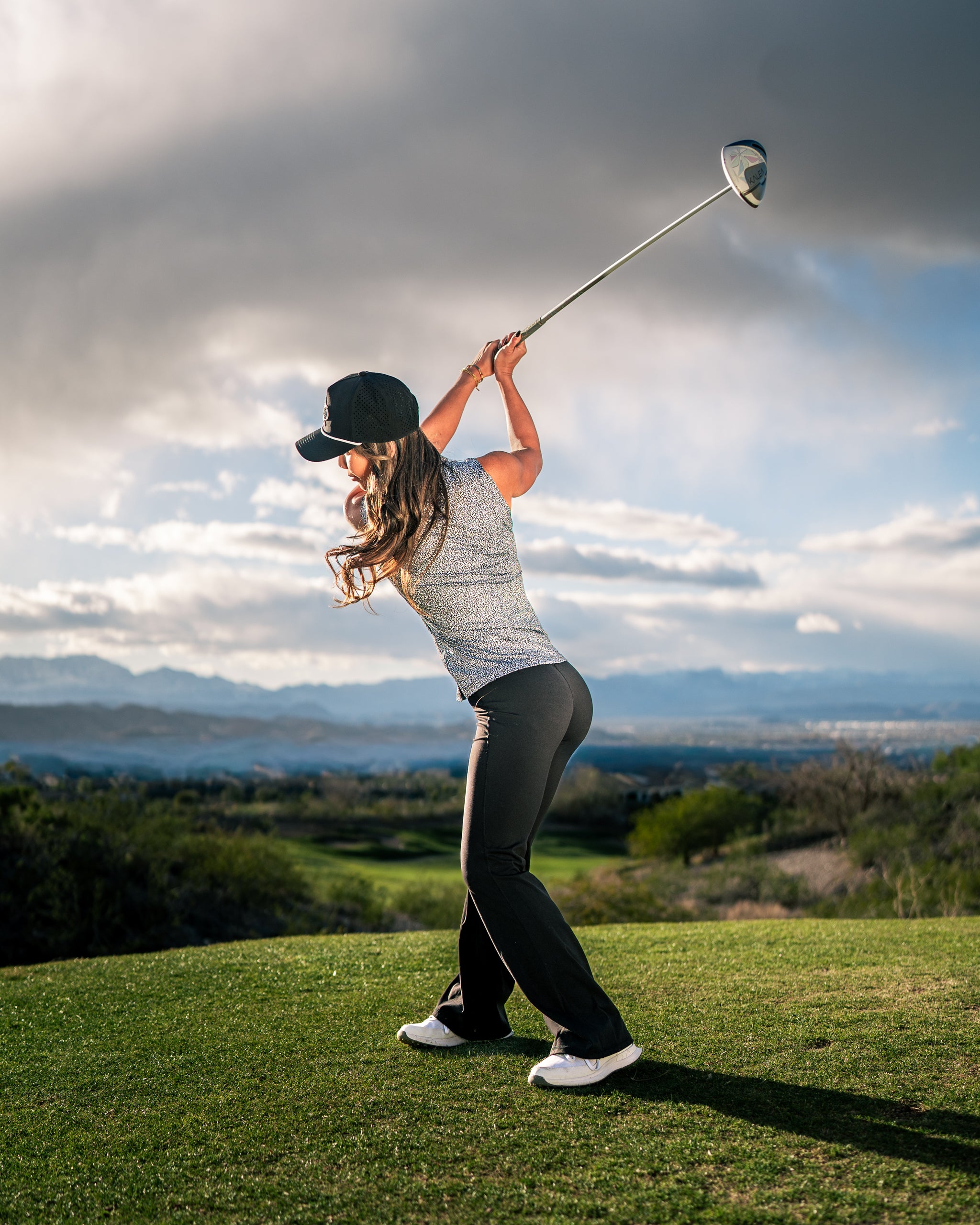 A woman in a stylish golf outfit swings a club on a scenic course, showcasing the Women's Coral Cascades apparel.